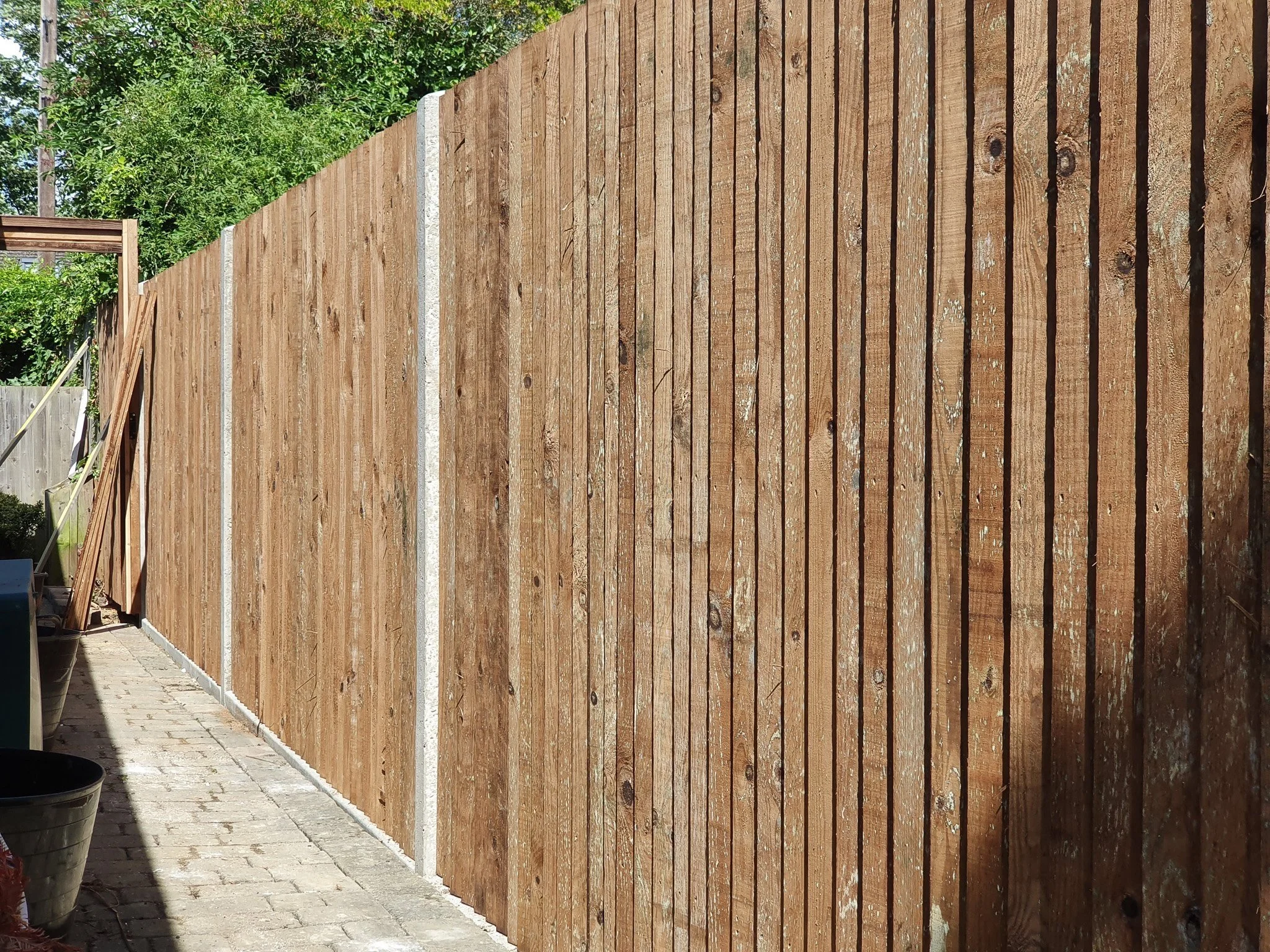 A newly installed wooden privacy fence along a stone-paved walkway with trees and greenery in the background.