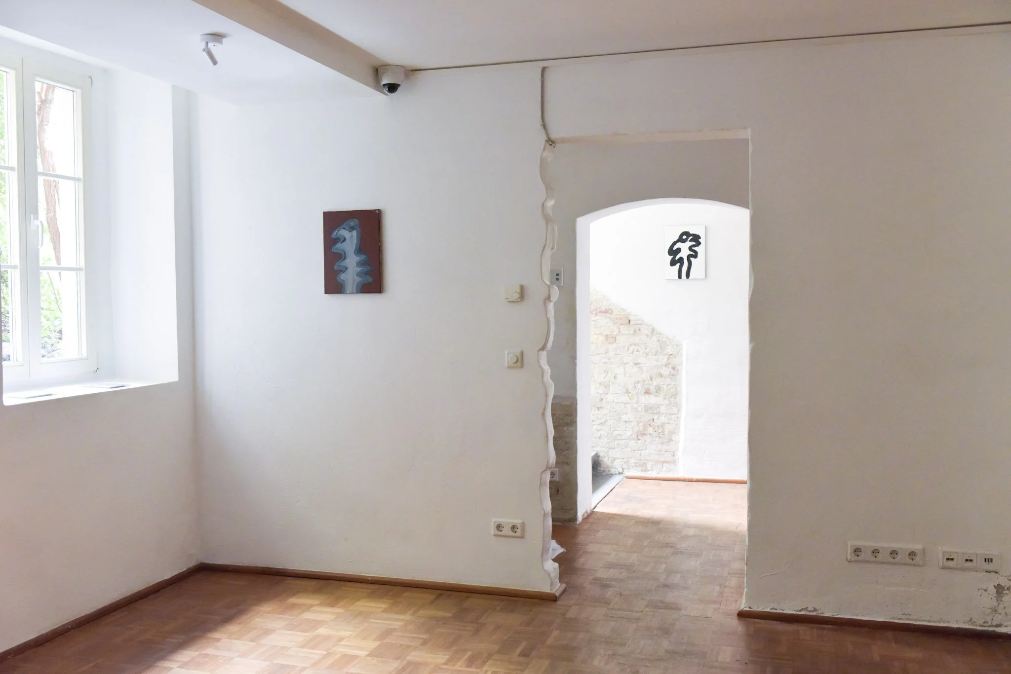 A corner of a bright room with white walls and a wooden floor. There is a window on the left side letting in natural light. Artwork with abstract designs hangs on the walls. An open doorway leads to another space with visible brickwork outside. Elect