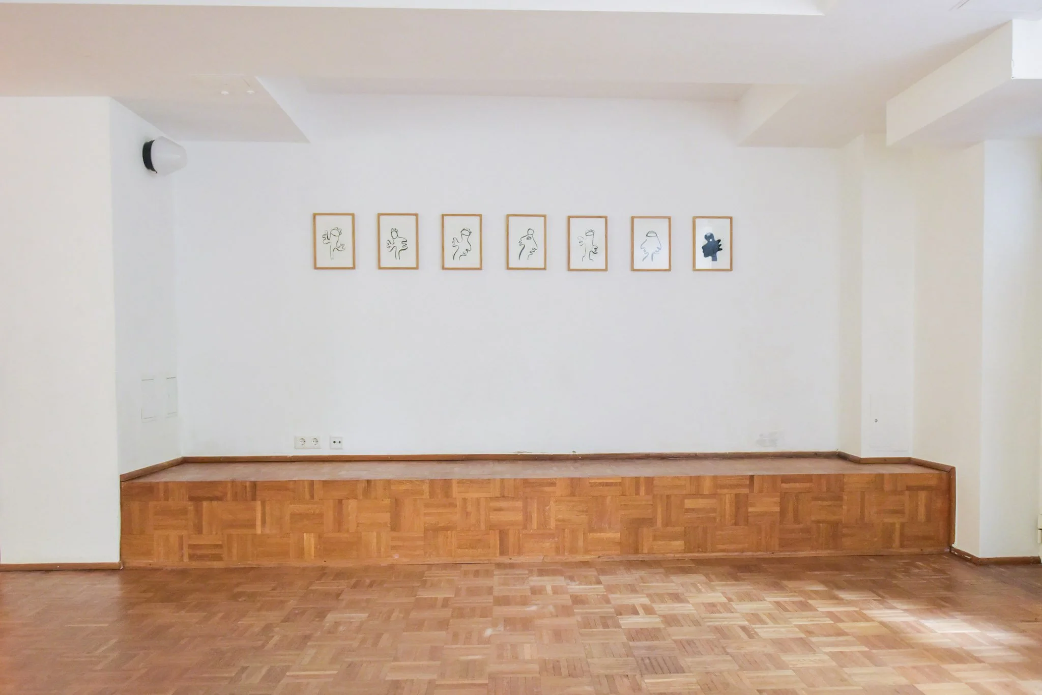 Empty art gallery with seven framed line drawings of abstract faces on a white wall, wooden floor, and a small stage.