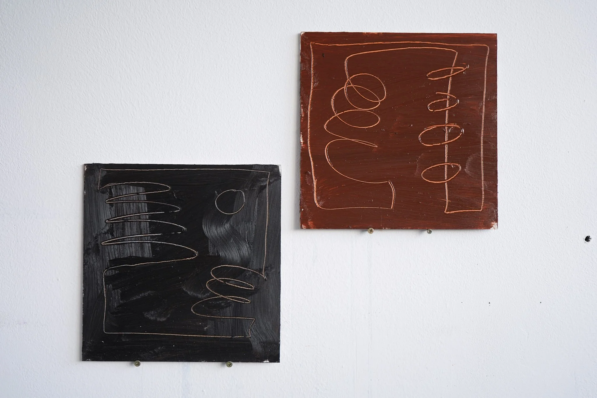 Two abstract paintings on a white wall, one black with a copper-colored spiral, and one reddish-brown with copper-colored swirls and lines.
