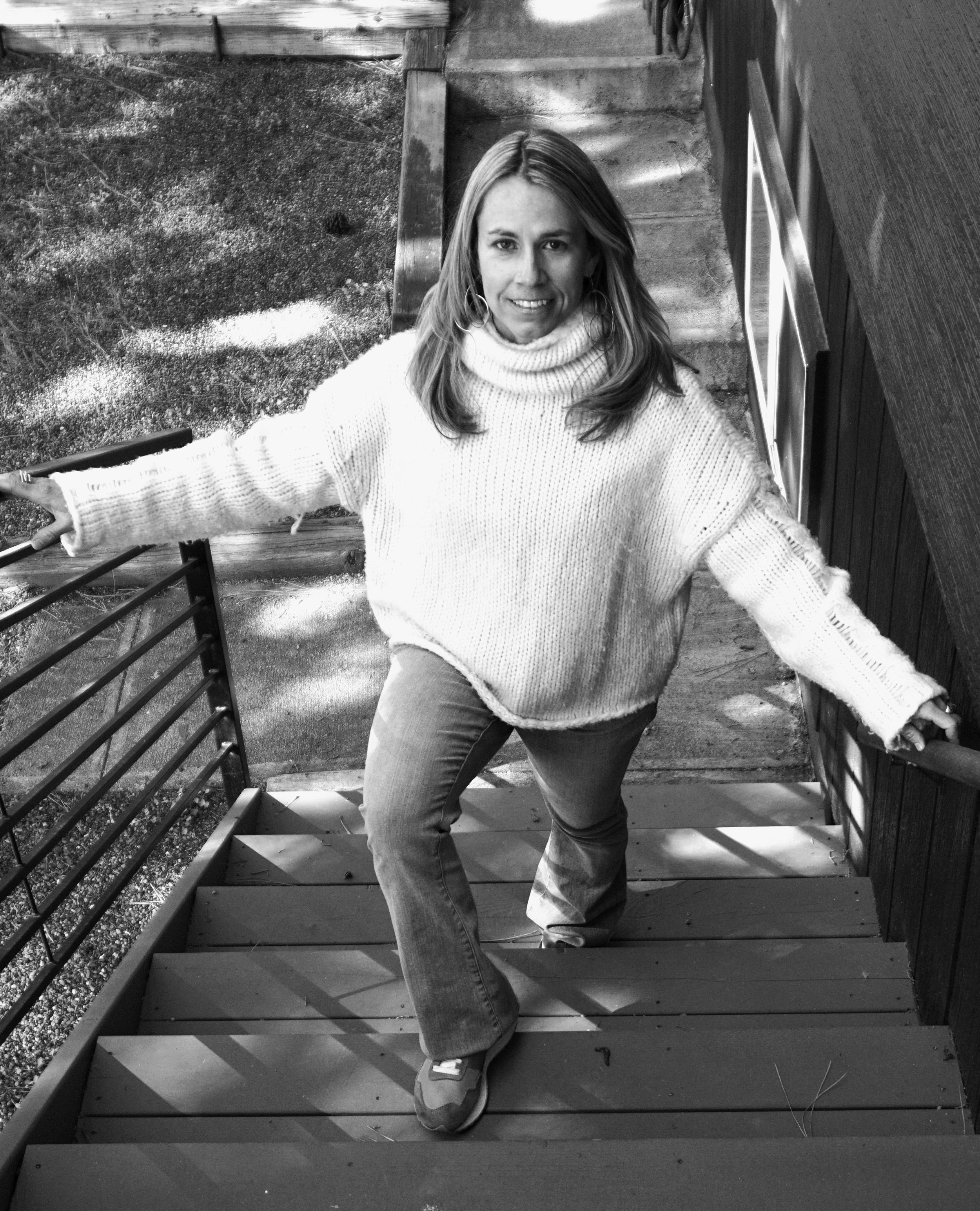 A woman with shoulder-length hair, wearing hoop earrings, a turtleneck sweater, jeans, and sneakers, posing on an outdoor wooden staircase with metal railings, smiling at the camera.