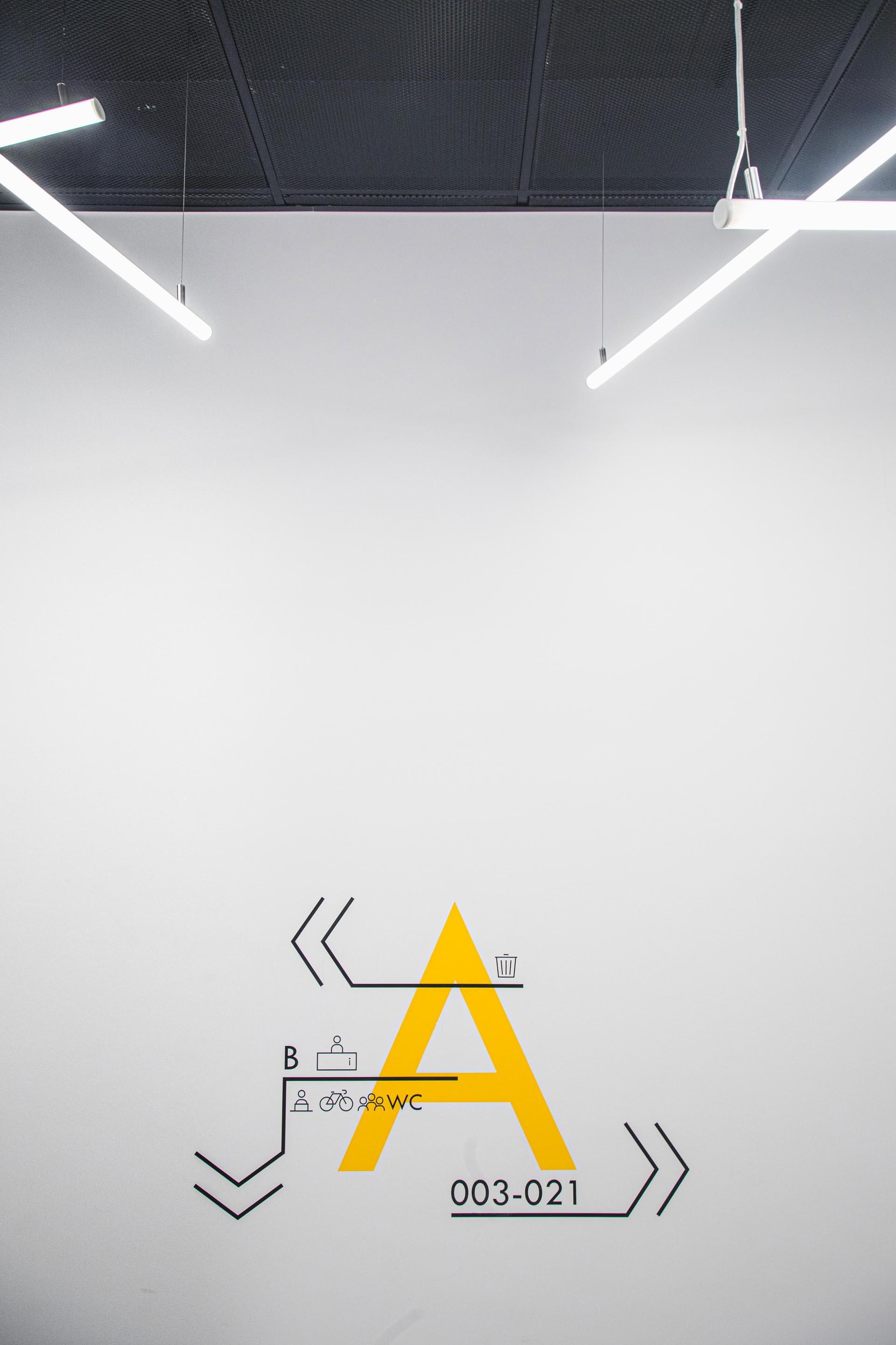 Interior of a modern building with hanging LED lights, featuring a wall with black and yellow graphic design resembling a schematic diagram with symbols, the letter 'A' prominently displayed in yellow, and room or office numbers.