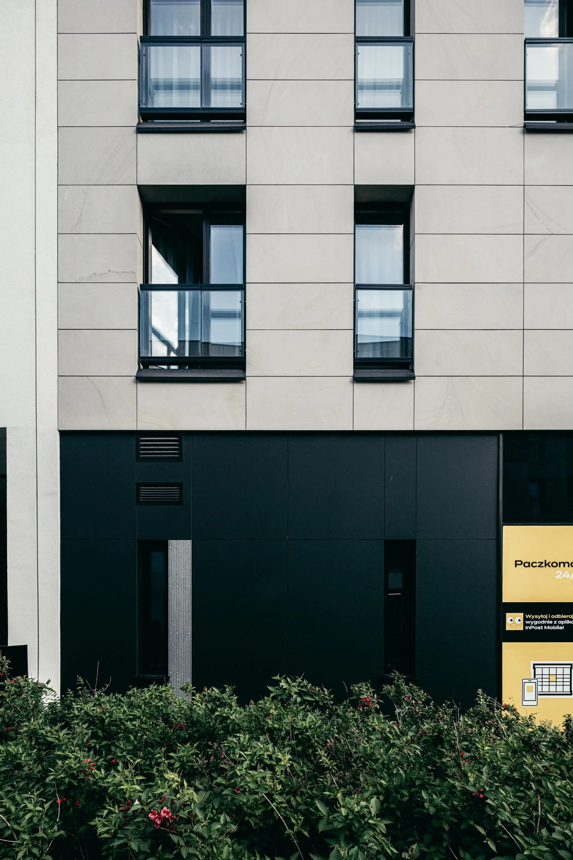 Modern building facade with beige and black panels, several windows with glass balconies, and greenery with red flowers at the bottom.