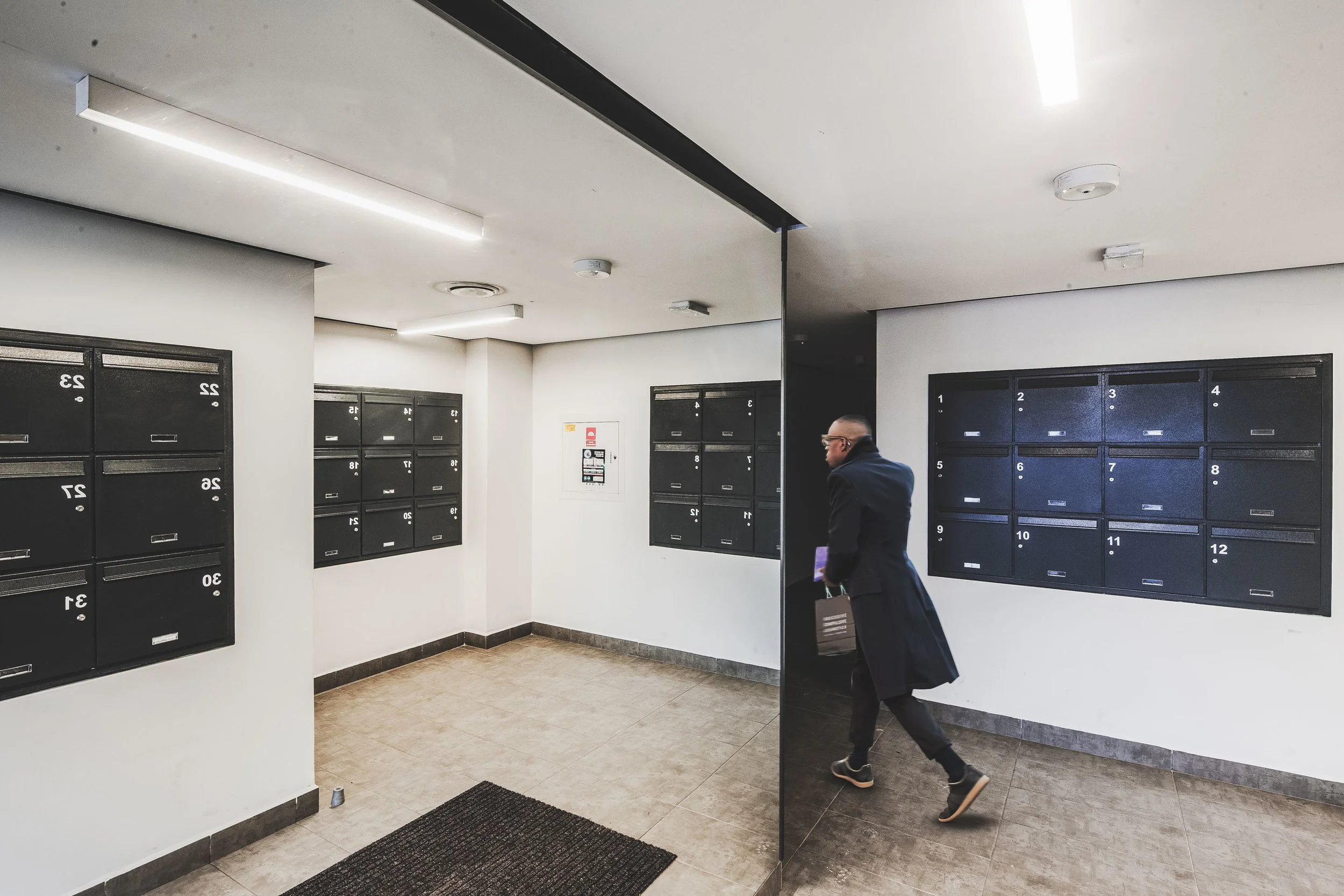 An indoor mailroom with black mailboxes arranged on white walls, and a person walking past a mirror dividing the room.