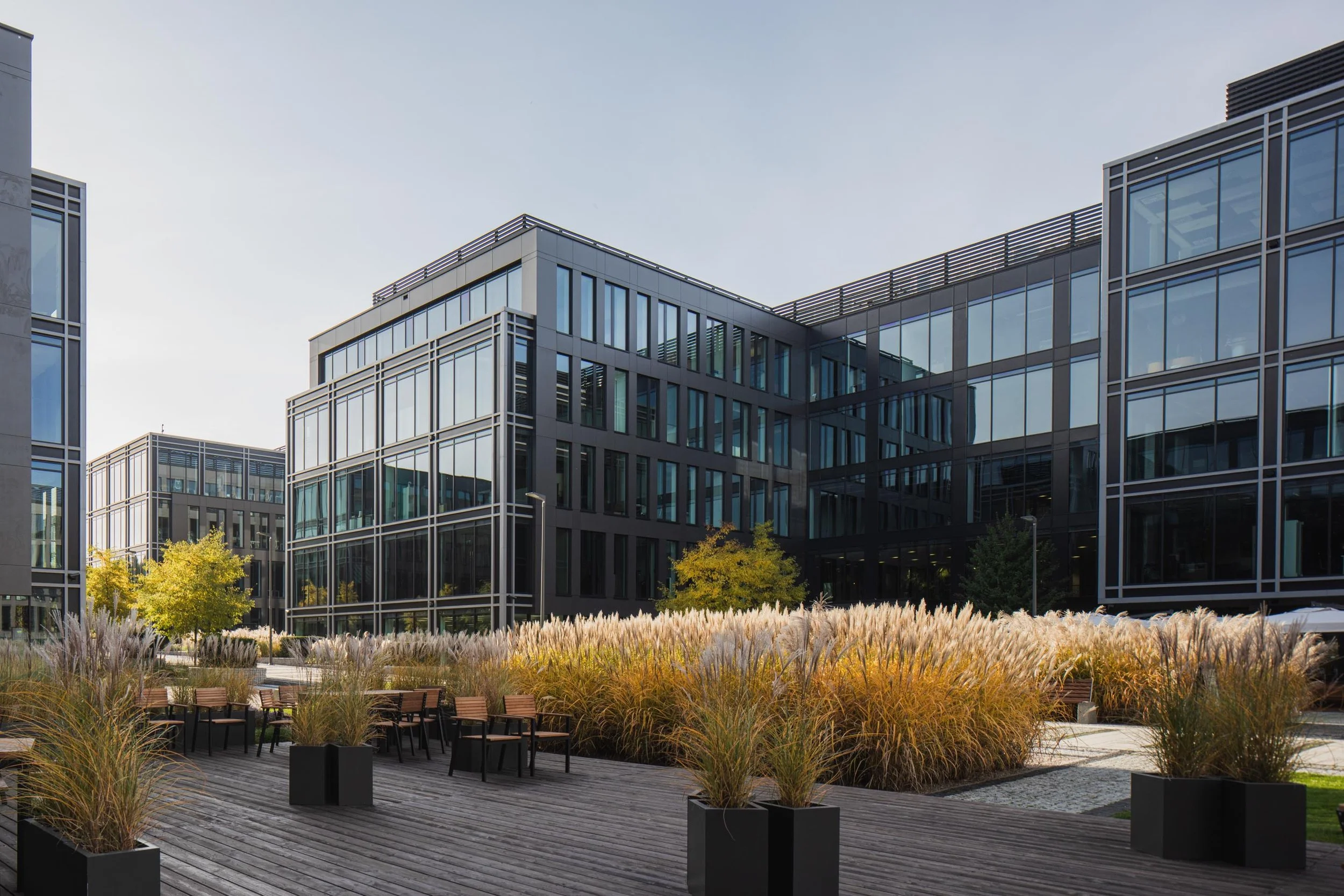 Modern office building with glass windows and surrounding outdoor seating area with plants and trees.