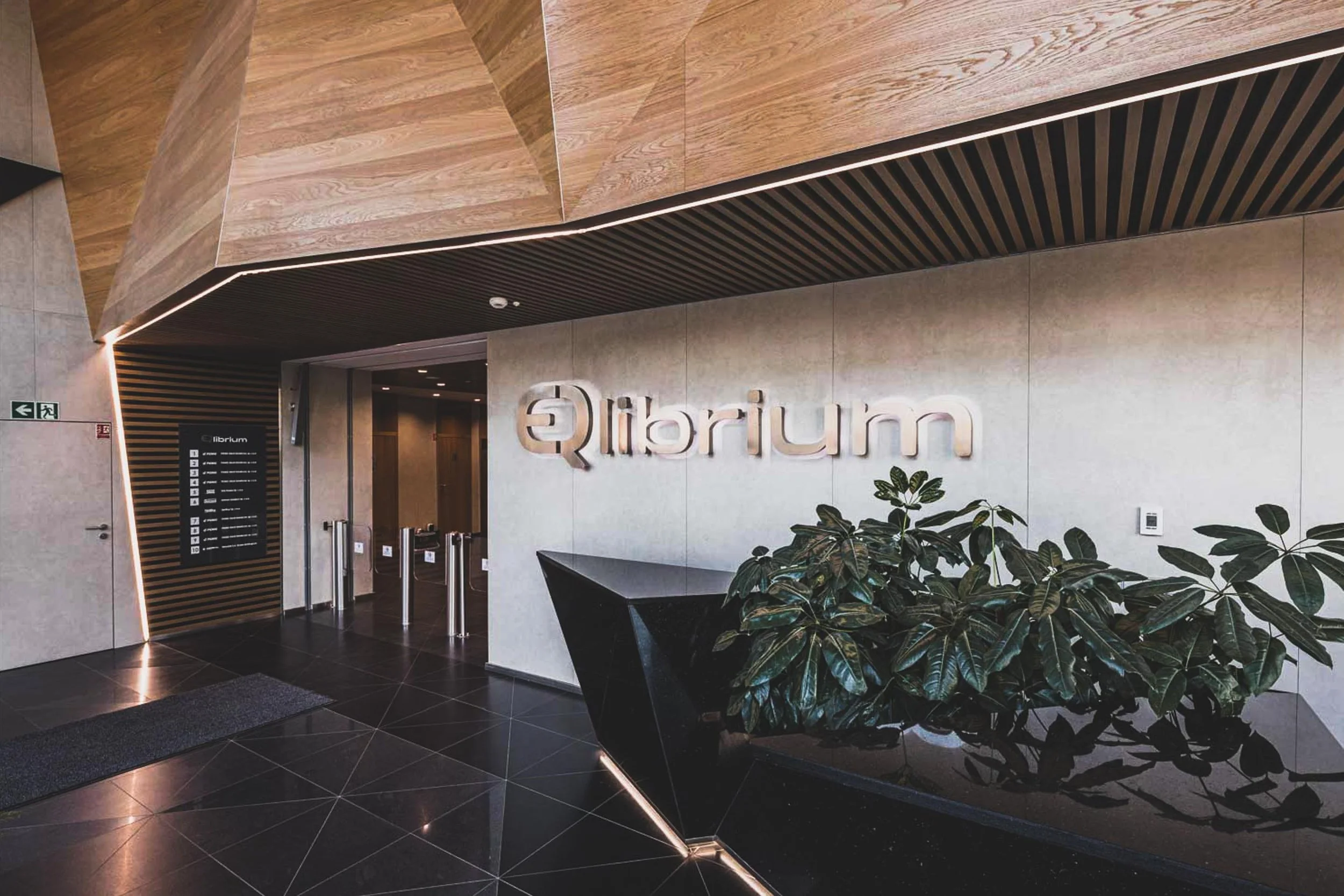 Modern lobby area with a large sign reading 'ilibrum' on the wall, a black geometric reception desk, a potted plant, and an electronic thermostat on the wall.