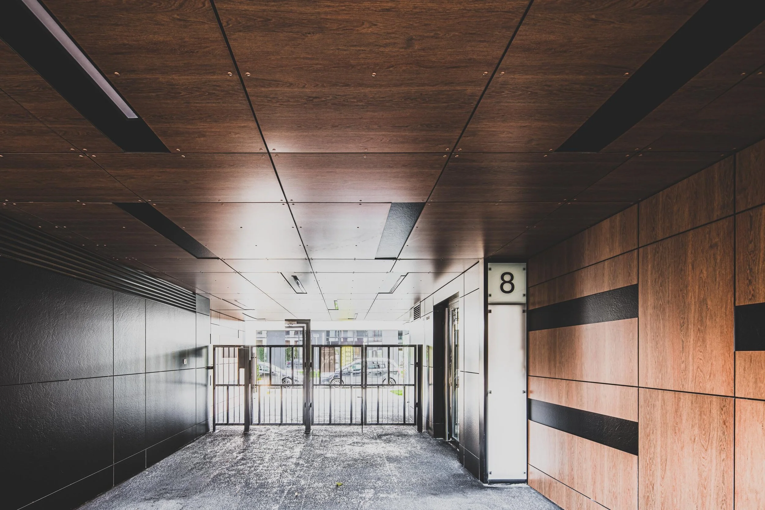 Interior of a modern building with wood-paneled ceiling and walls, black accents, and an elevator with a sign showing the number 8, near glass doors leading outside.