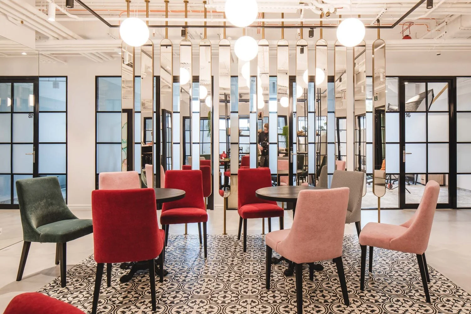 Modern restaurant or cafe with colorful upholstered chairs around black tables, glass partition with black frames, pendant lighting, patterned area rug, and people in the background.