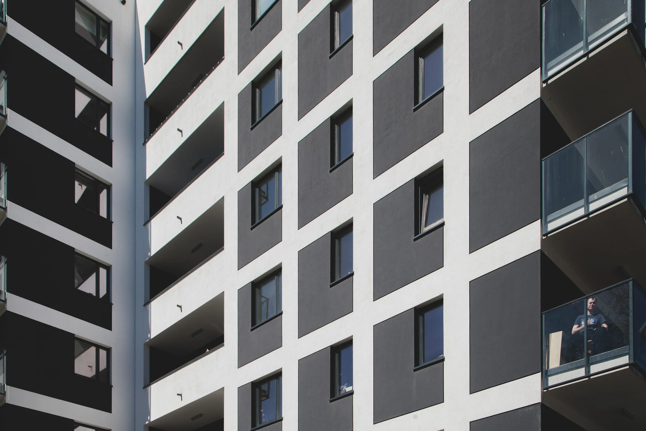 Close-up of a modern apartment building with multiple balconies and windows, some balconies have glass railings, one person is visible standing on a balcony.