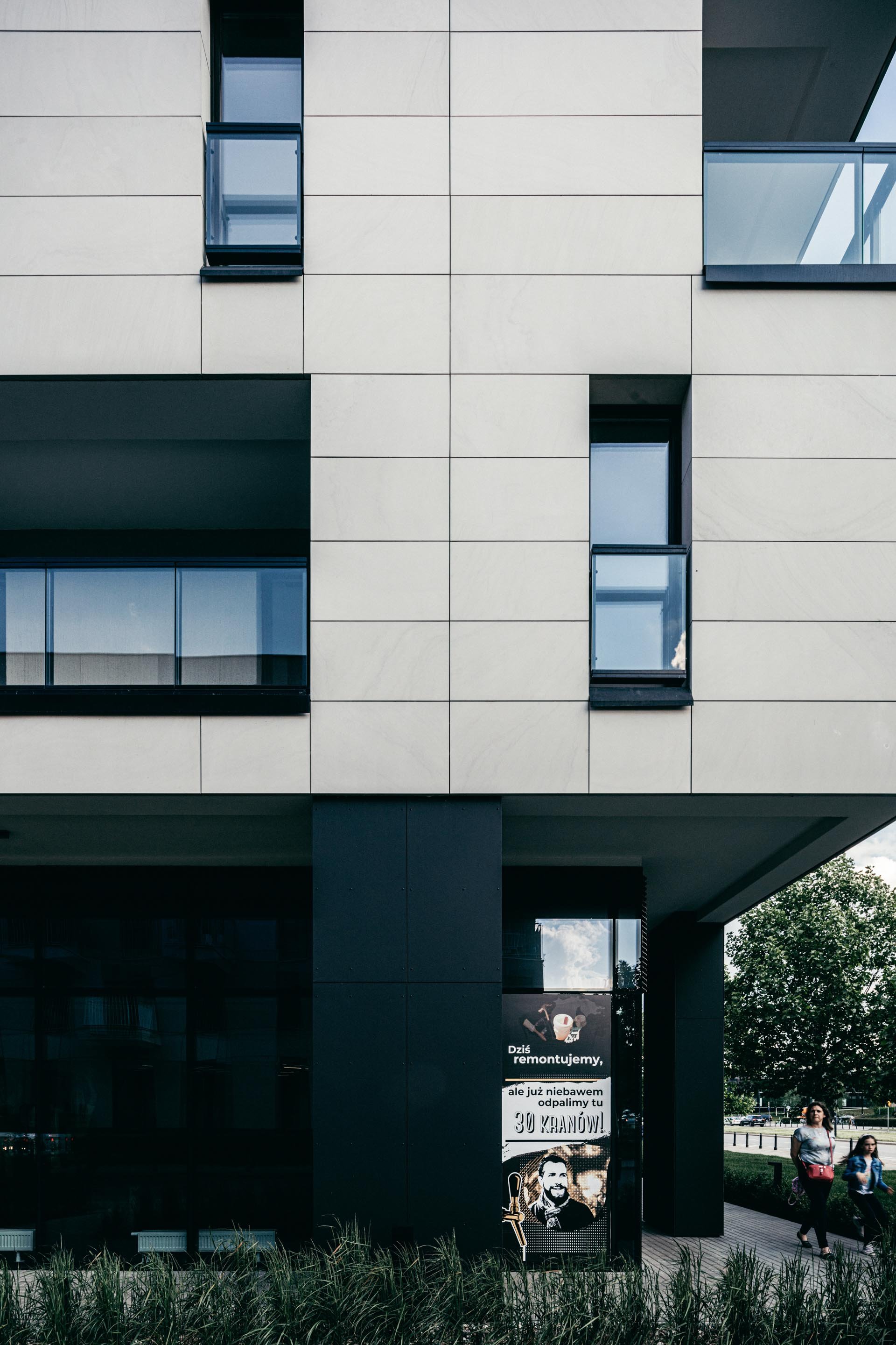 Modern building with beige and black exterior walls, multiple rectangular windows, and a sign advertising renovation work with an illustrated man wearing a suit and holding a pitchfork, and people walking outside.