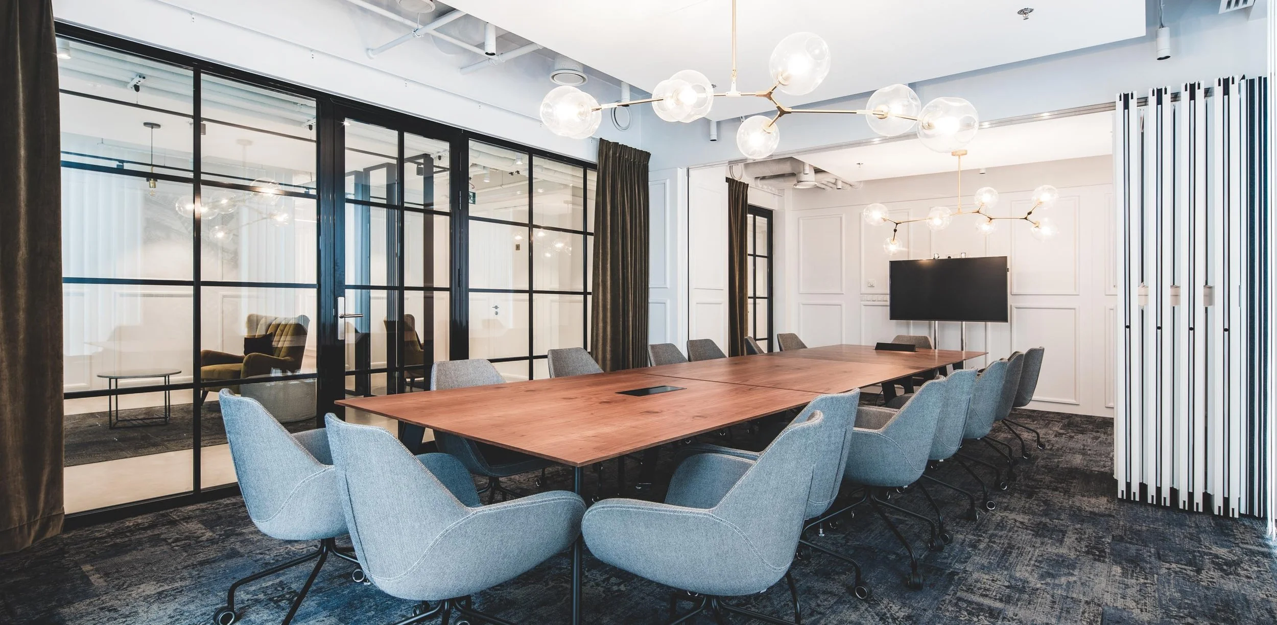 Modern conference room with a large wooden table surrounded by gray office chairs, decorative lighting fixtures overhead, and a large flat-screen TV on the white paneled wall. Glass-walled private rooms are visible in the background.