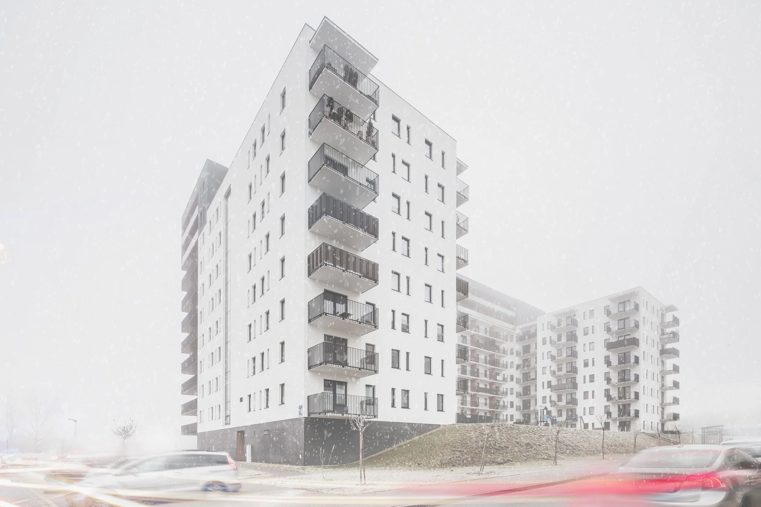 Modern white apartment buildings with balconies, snow falling, cars passing by, and a small grassy hill in the foreground.