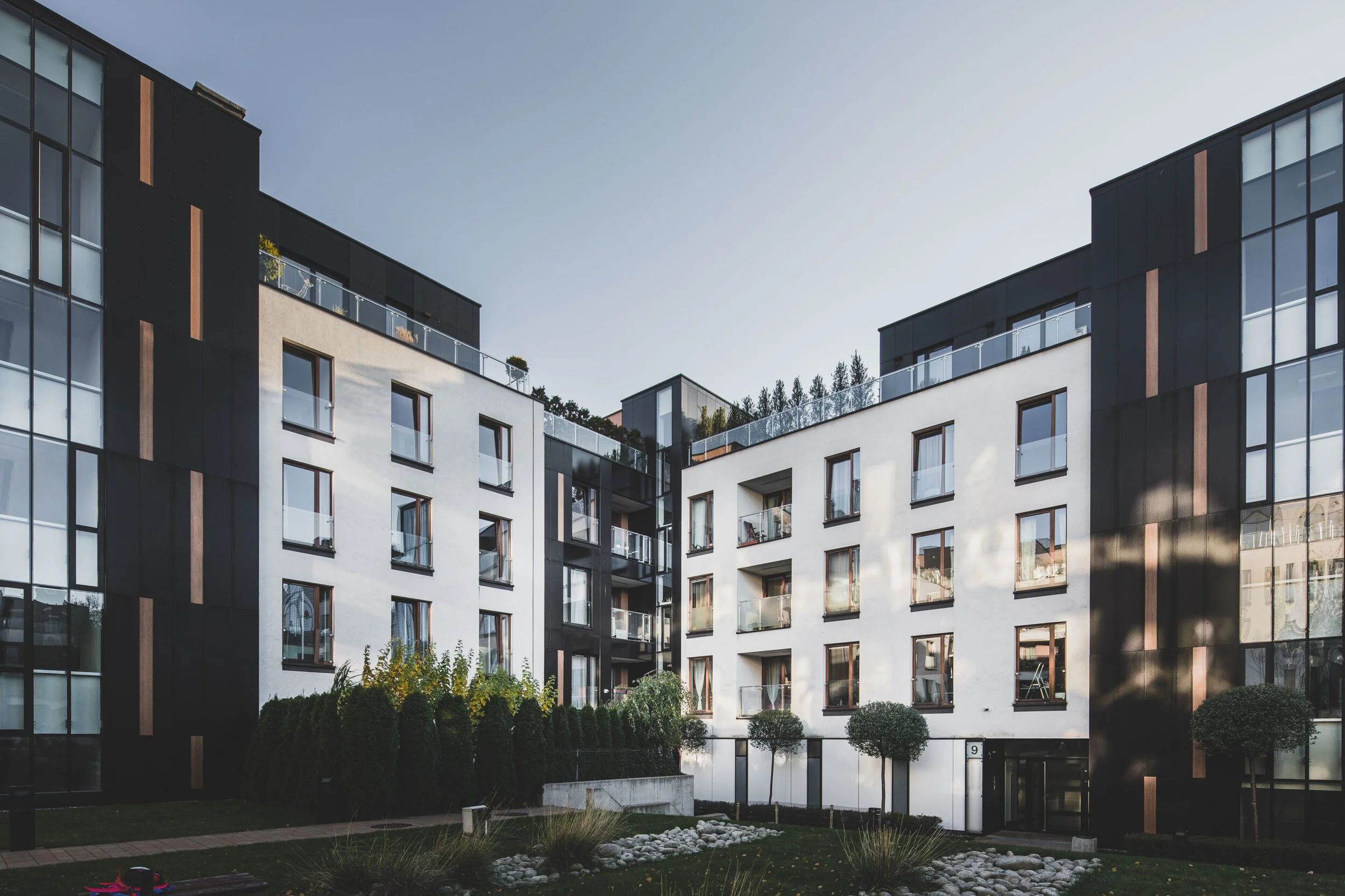 Modern apartment building with black and white facade, glass balconies, and landscaped courtyard with trees and rocks.