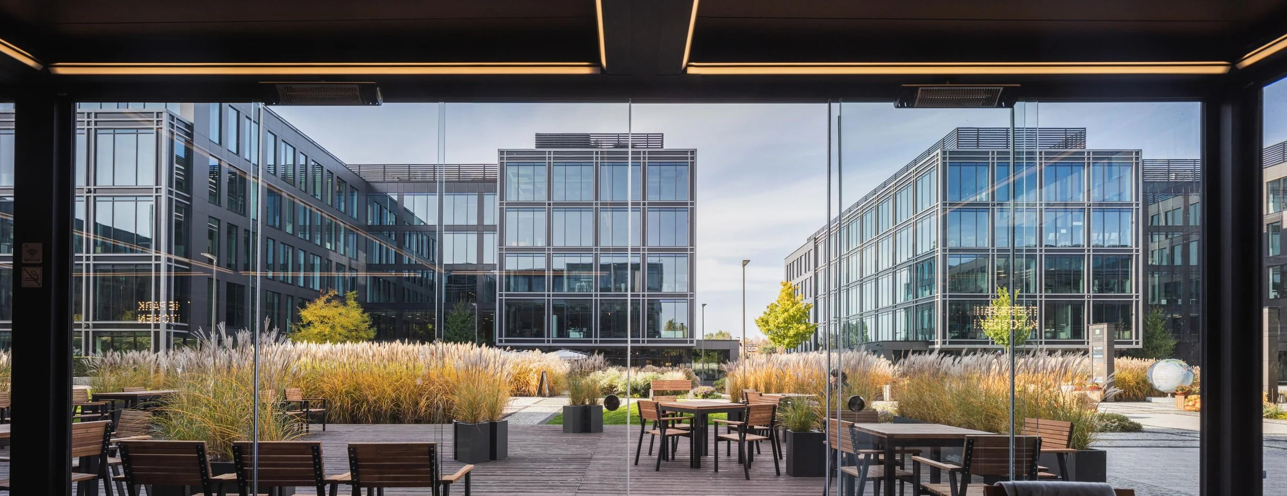 Modern office buildings with glass facades seen from inside a workspace with wooden tables and chairs and landscaped outdoor area