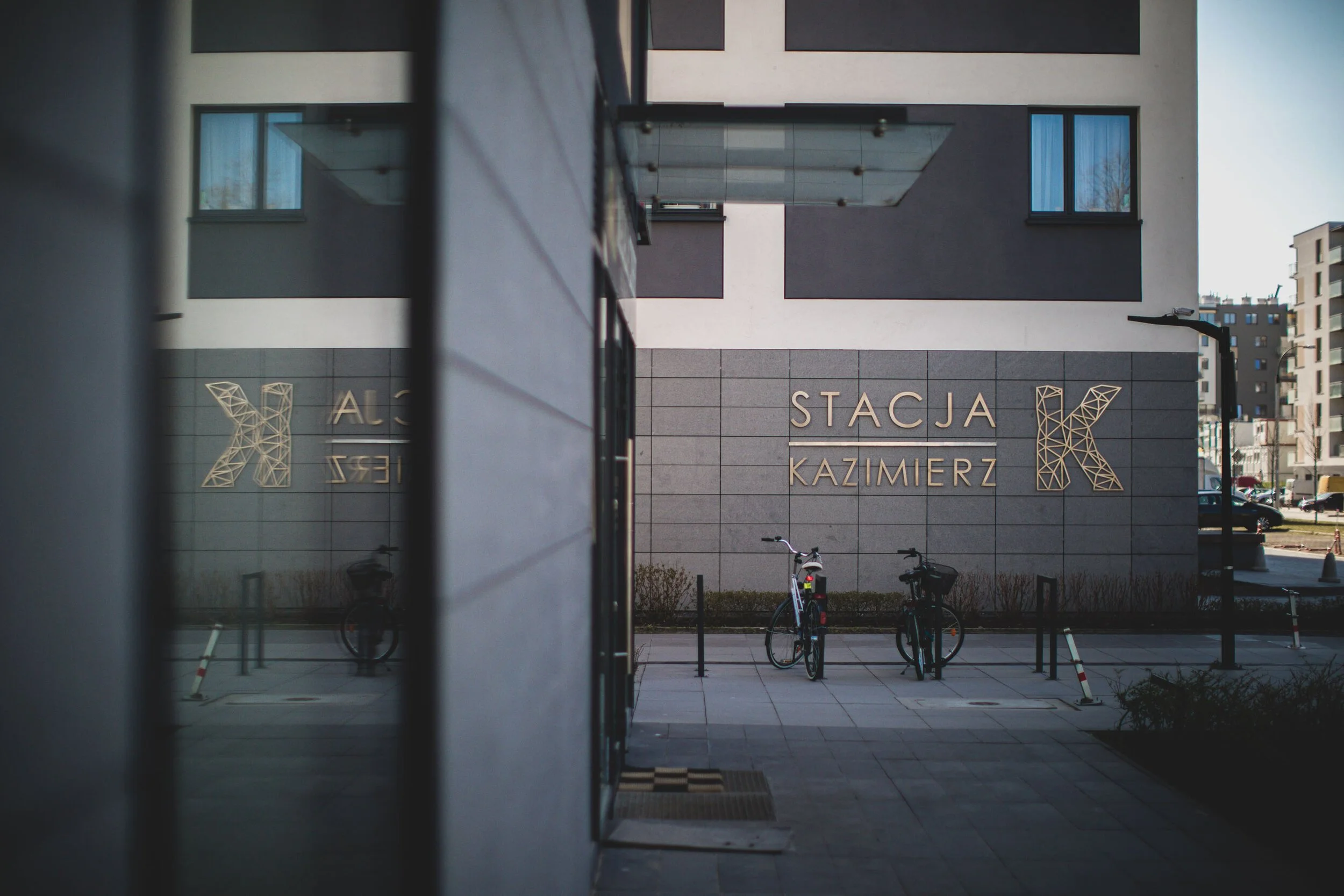 View of a modern building with the sign 'Stacja Kazimierz' on the wall, two bicycles parked outside, and other buildings in the background.