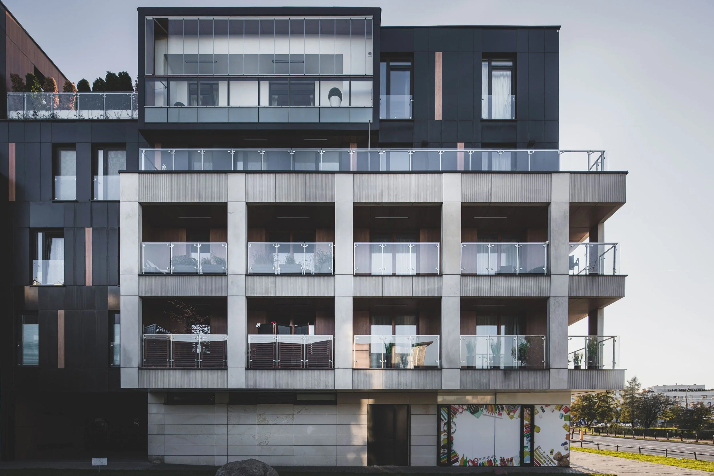 Modern multi-story apartment building with balconies that have glass railings and large windows, situated in an urban area with trees and a street nearby.