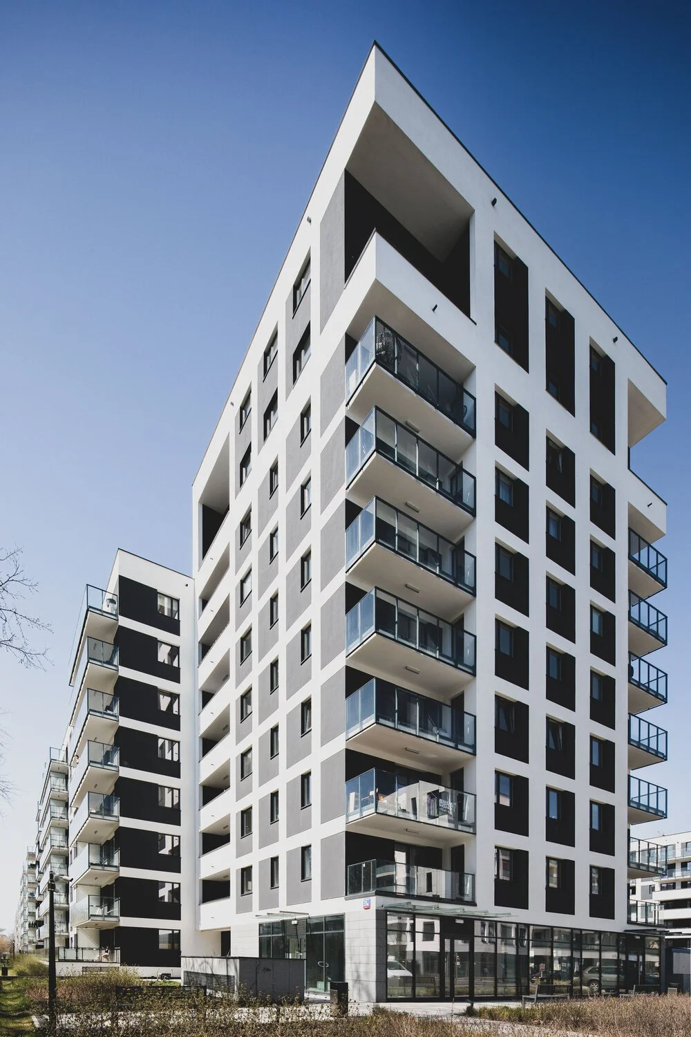 Modern white multi-story apartment building with balconies and black window frames, under a clear blue sky.