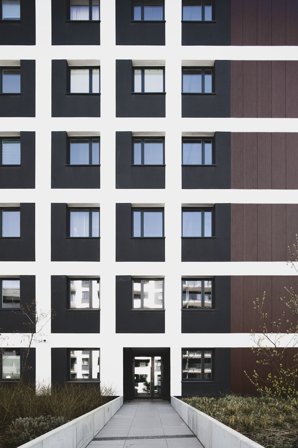 Front view of a modern apartment building with black and white exterior, multiple windows, and a pathway leading to the entrance.