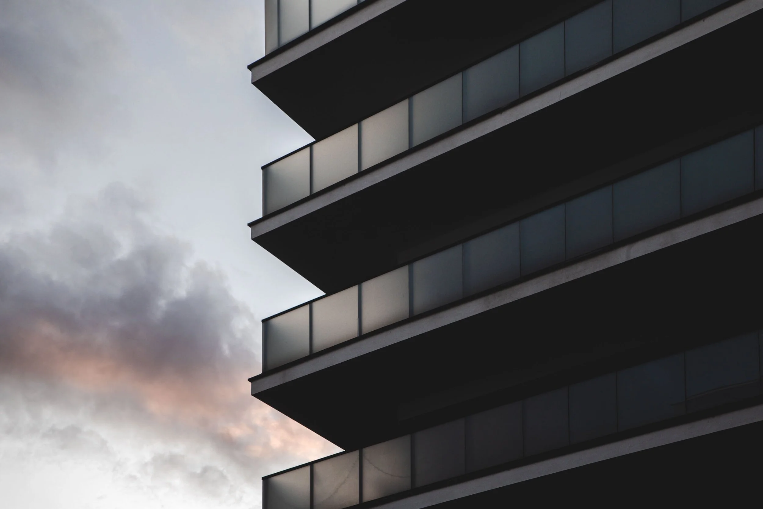 A modern multi-story building with glass balconies against a cloudy sky at sunset.