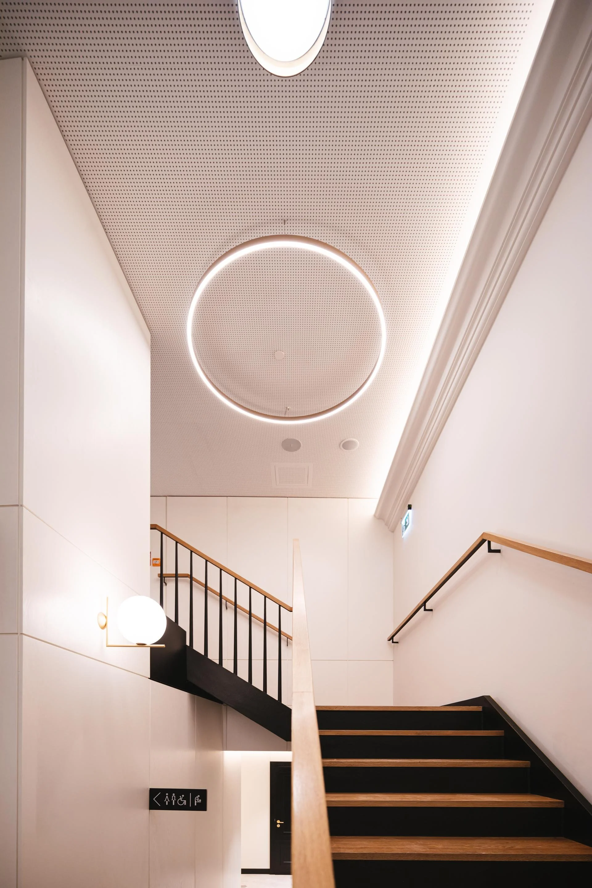 Interior view of a modern building with a staircase, wooden handrails, black steps with wood accents, white walls, and ceiling lighting fixtures, including a circular light feature and a globe-shaped wall lamp.