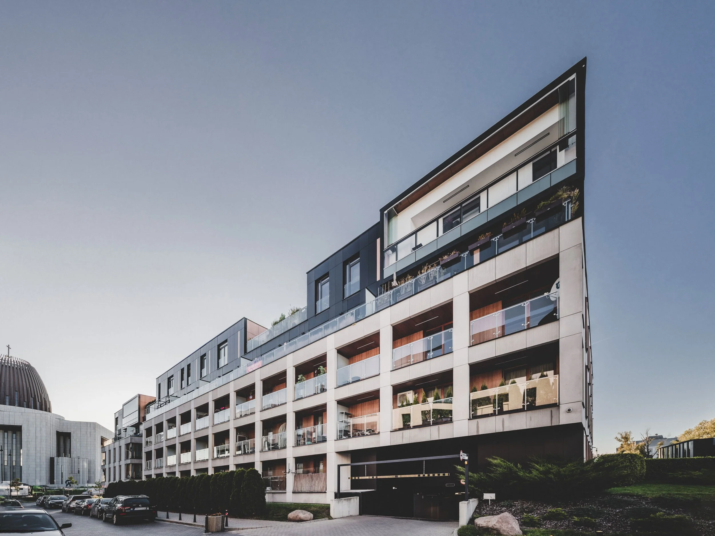 Modern multi-story residential building with balconies and glass railings, surrounded by parked cars and landscaping under a clear sky.