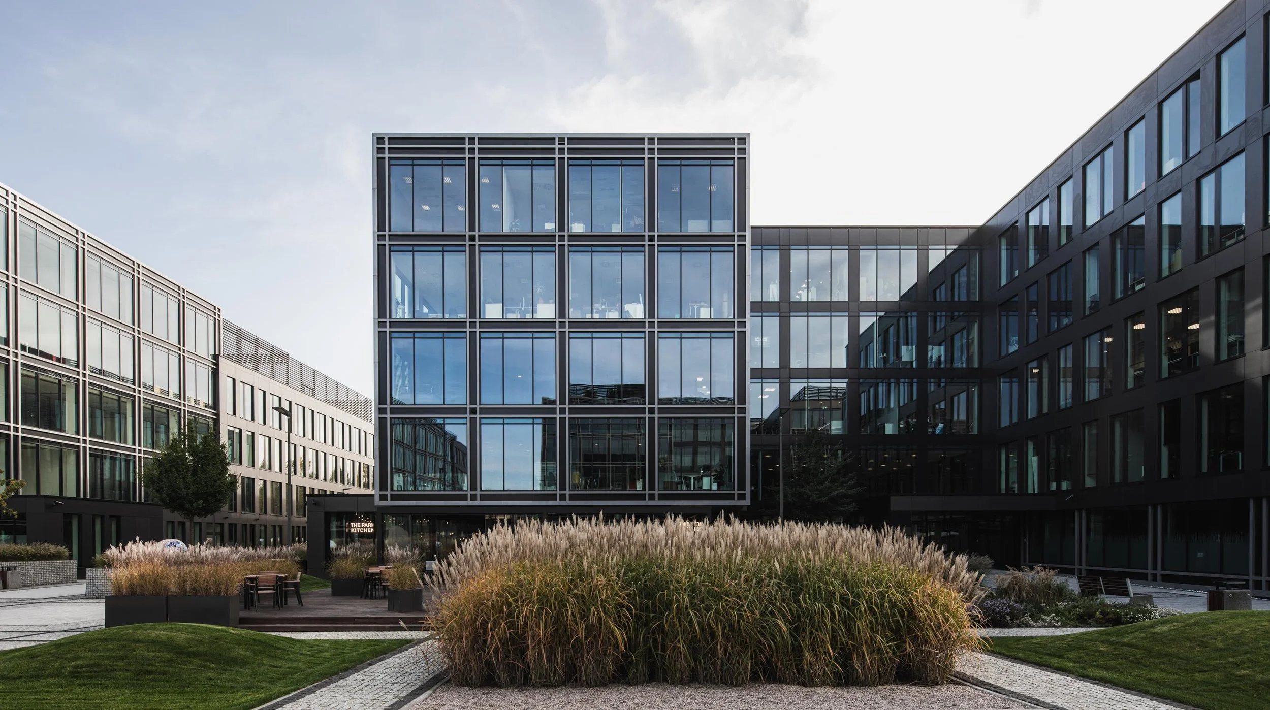Modern office building with glass windows, surrounded by landscaped garden with ornamental grasses and walking paths.