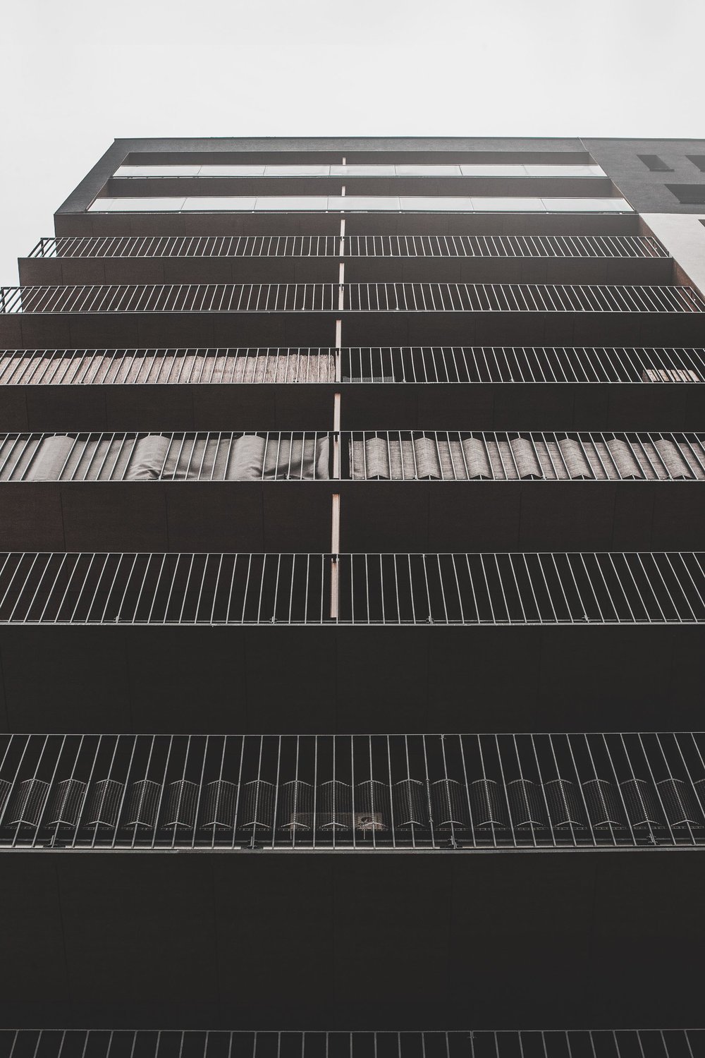 Low-angle view of a modern multi-story building with railings on each balcony and curtains visible through the windows, against a gray sky.