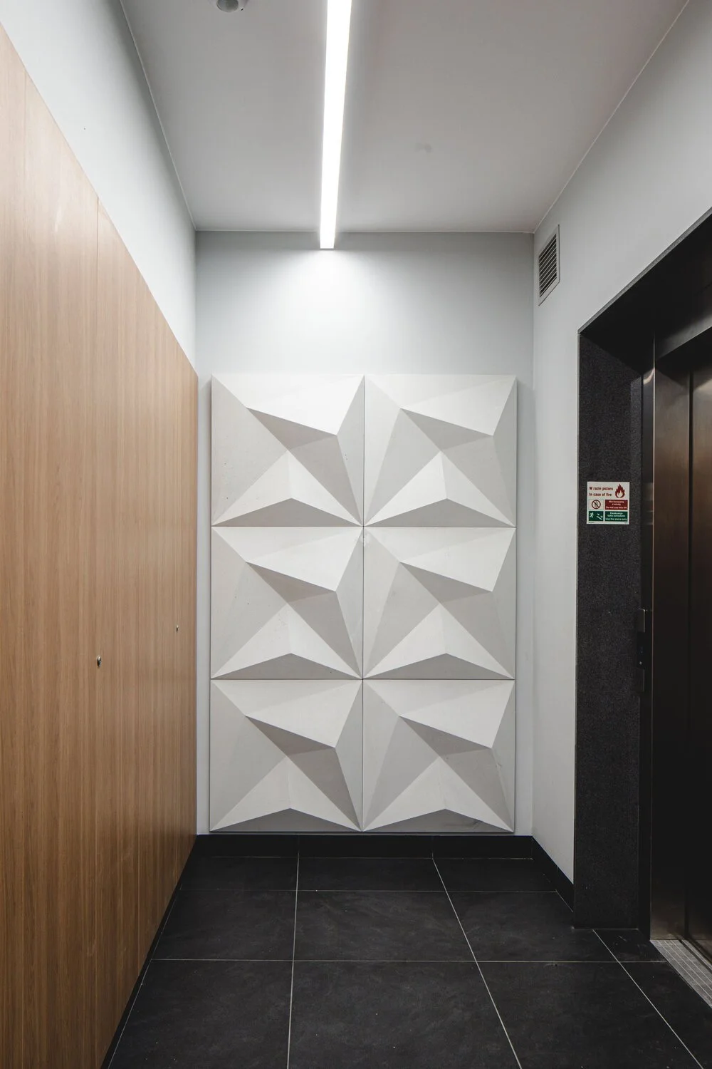 Interior wall with white three-dimensional geometric panels, wooden wall on the left, black tiles on the floor, and an elevator door on the right.