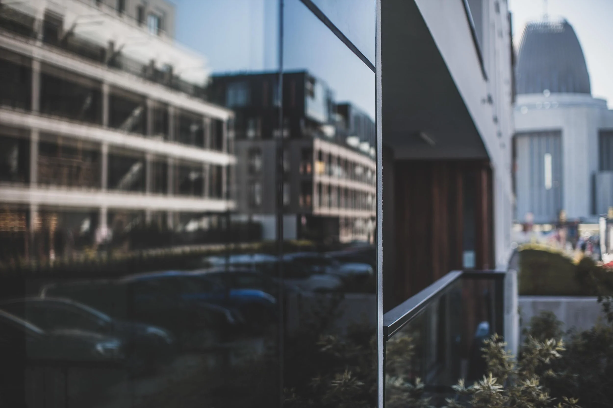 Reflection of modern buildings and parked cars on a glossy exterior wall of a building, with a blurred cityscape in the background.