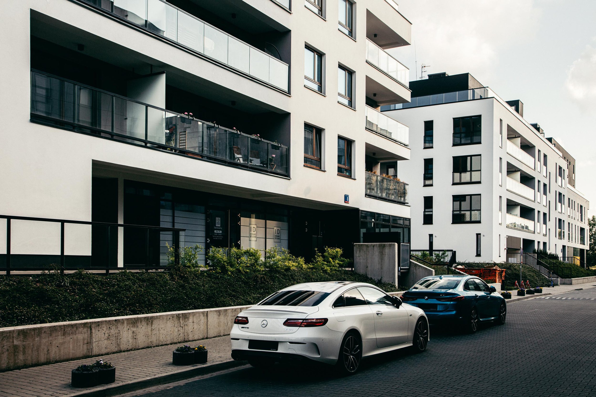 Modern white apartment building with balconies, parked white and blue cars, and small planters along the sidewalk.