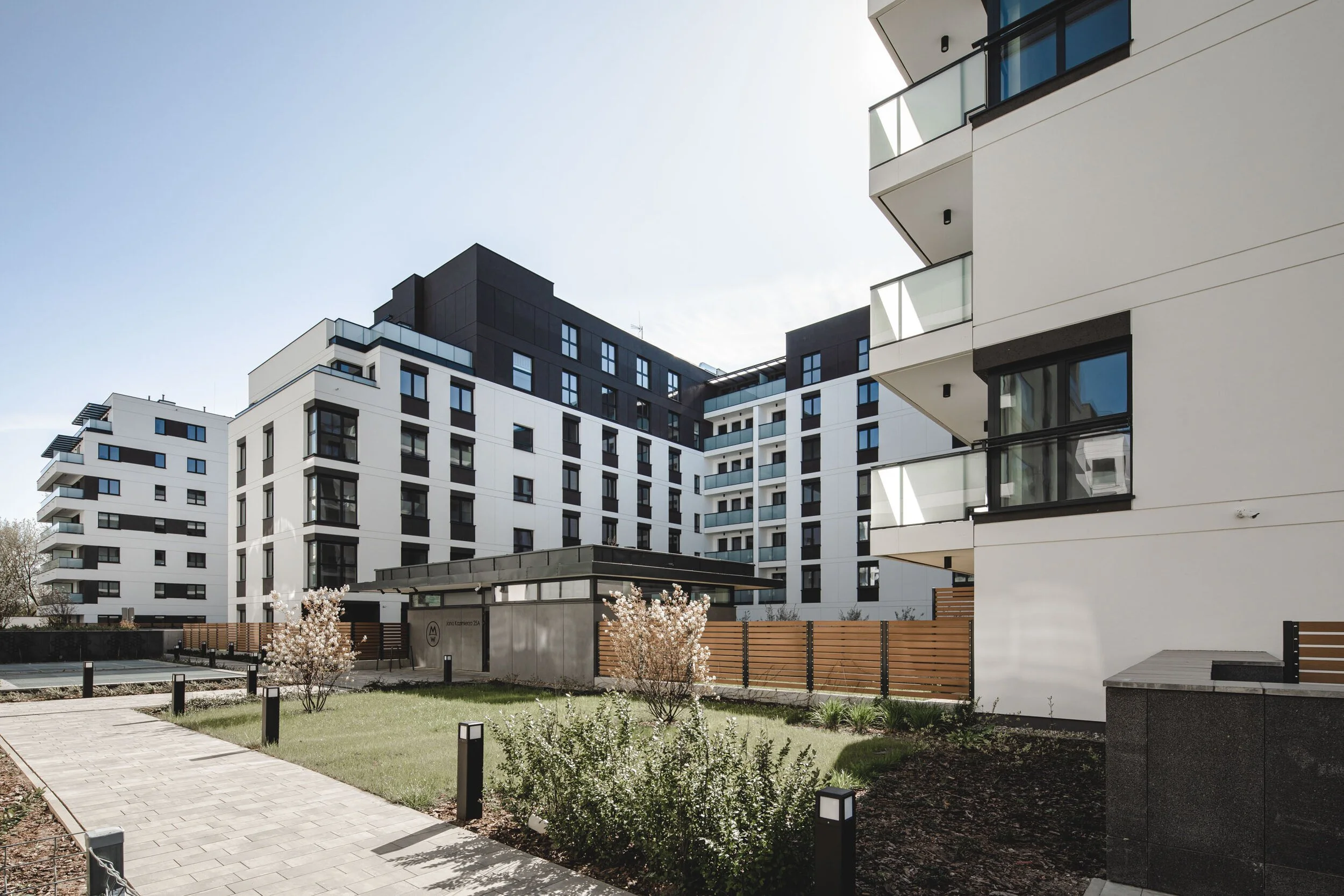Modern apartment complex with white and black facade, balconies with glass railings, well-maintained lawn, small flowering trees, paved walkway, and outdoor light fixtures.