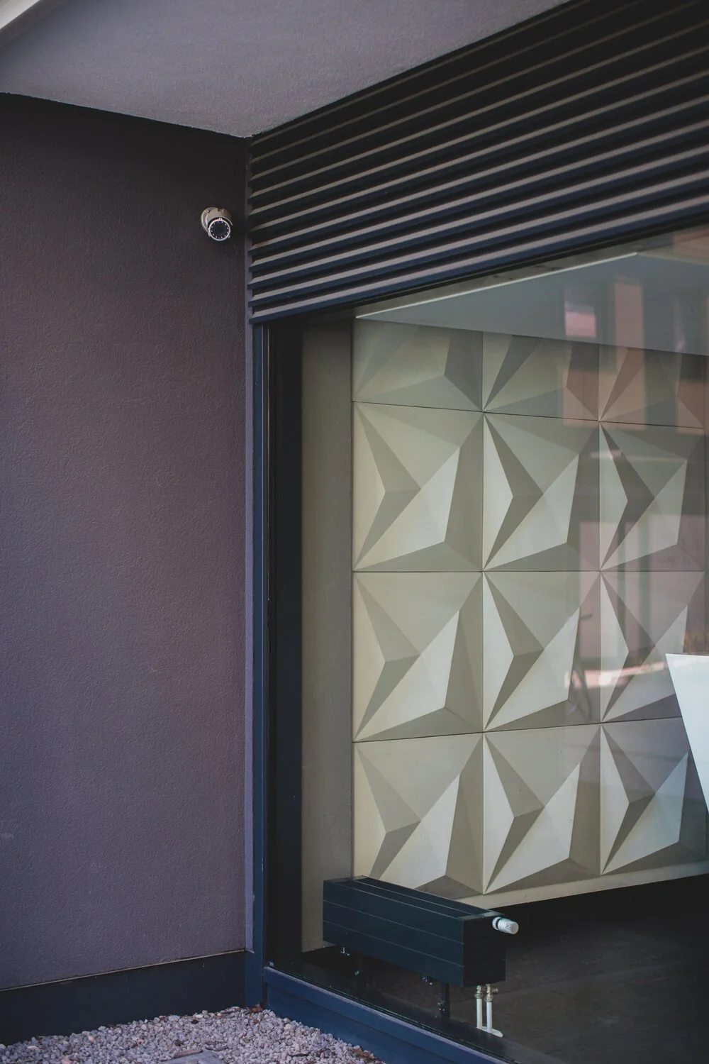 Security camera mounted in the upper corner of an exterior wall of a modern building with geometric patterned tiles visible through a glass window.