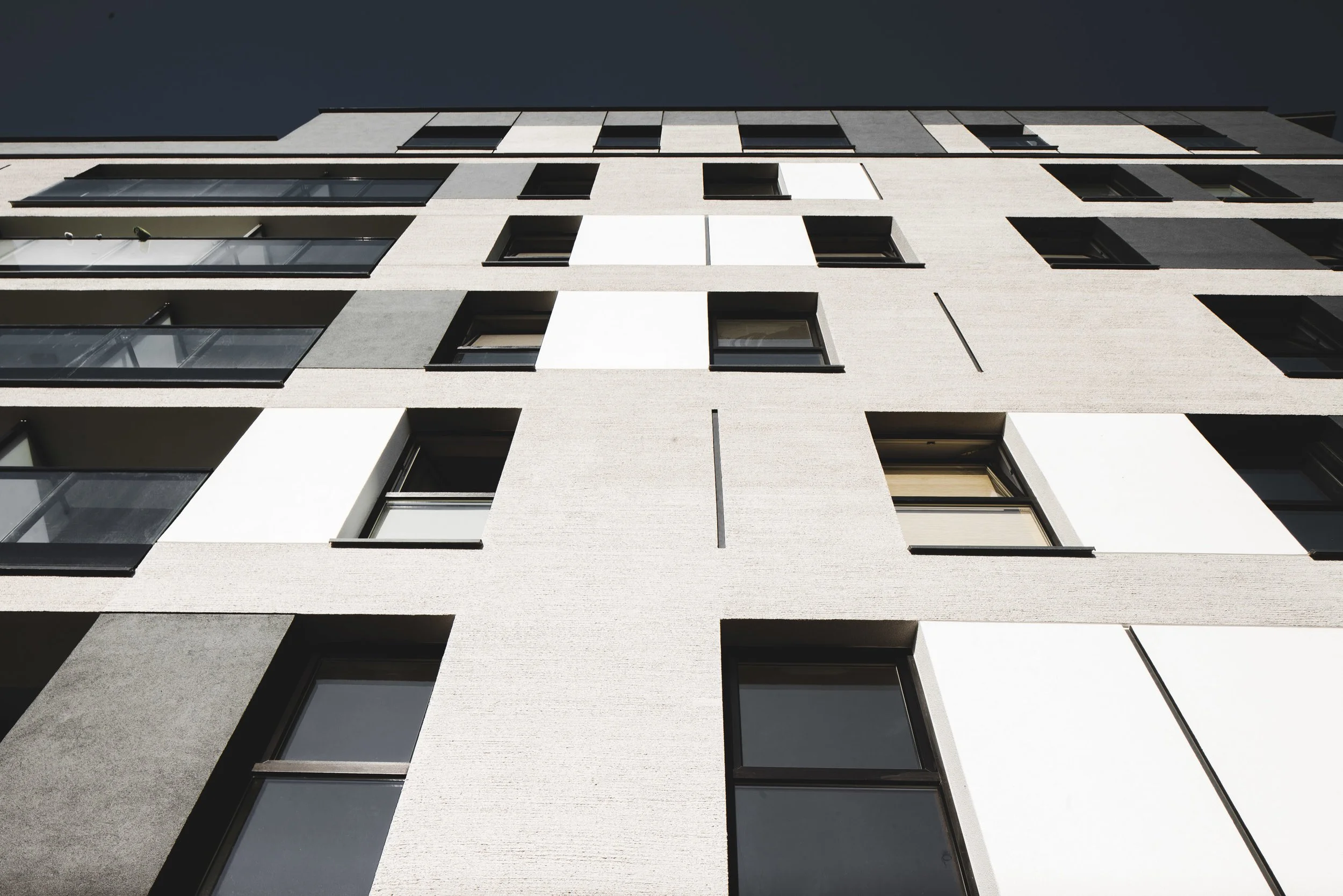 Low-angle view of a modern multi-story apartment building with white and gray facade, black-framed windows, and balconies under a dark sky.