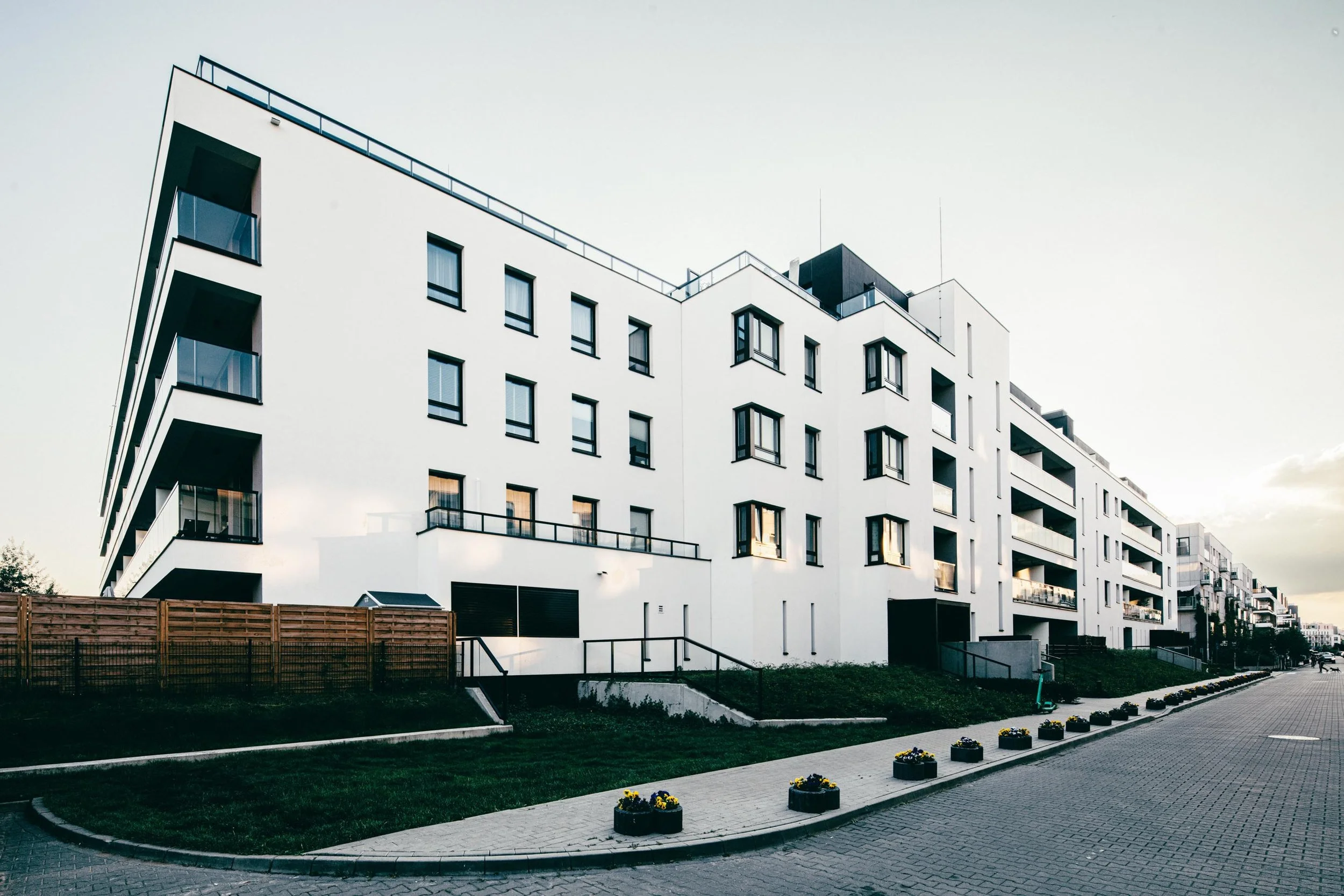 Modern white multi-story apartment building with balconies and black window frames, along a paved walkway with flower planters.