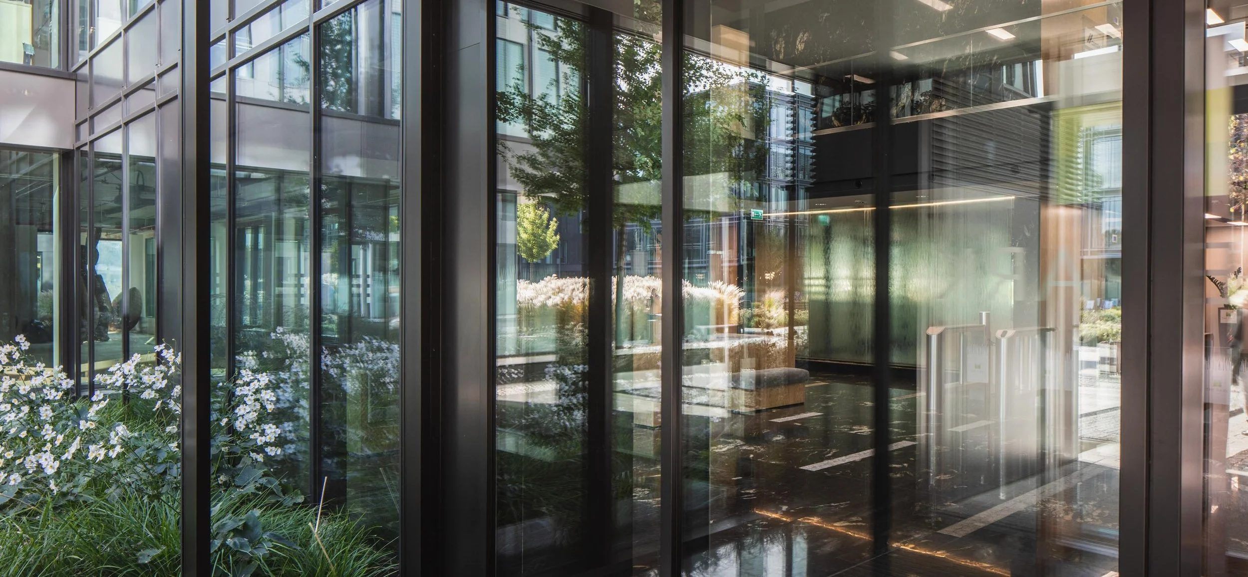 Glass exterior of a modern building with reflections of trees and a parking lot, including sliding doors at the entrance.
