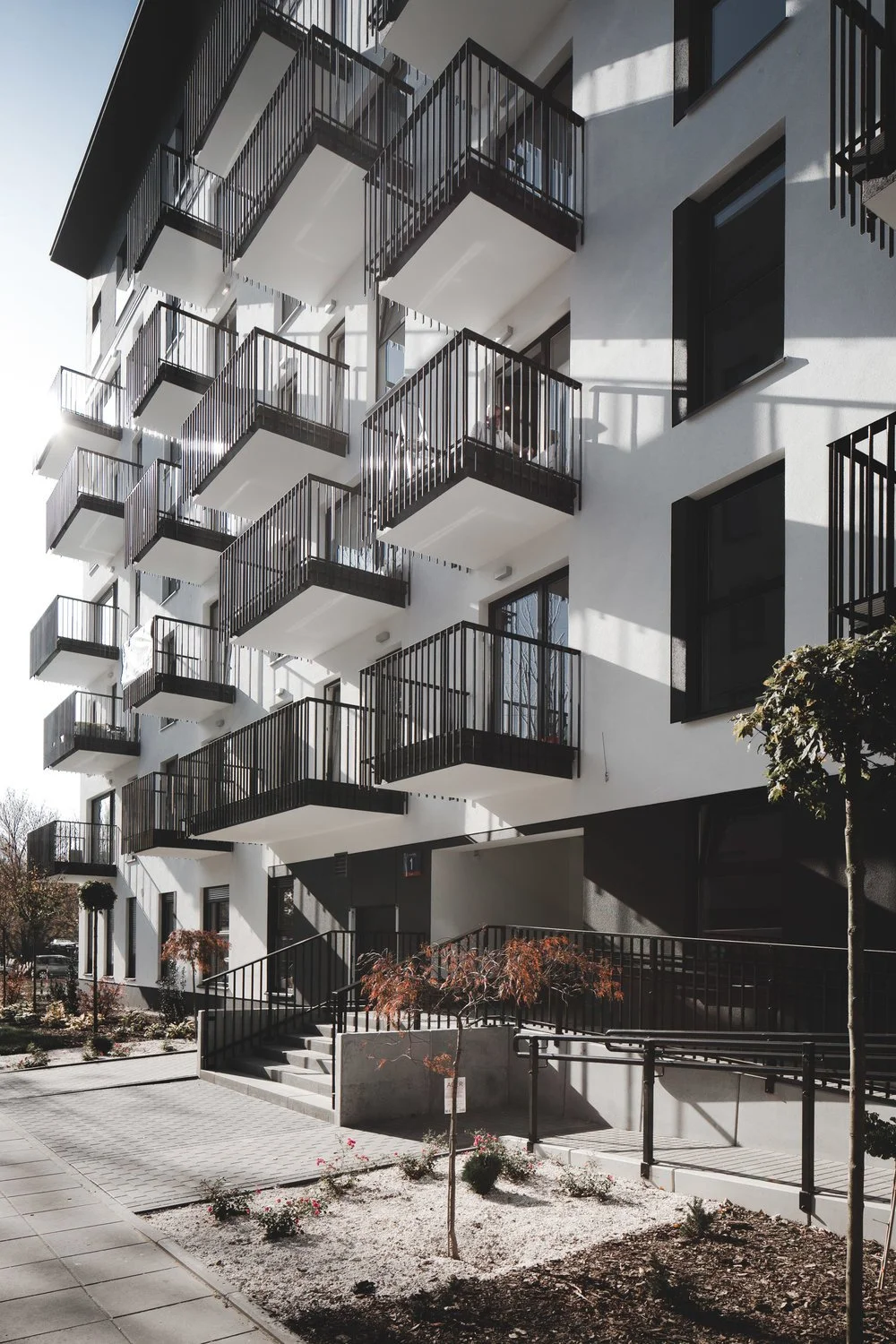 Modern white apartment building with multiple black metal balconies and large windows, sun casting shadows, landscaped area with small trees and flower beds in foreground.