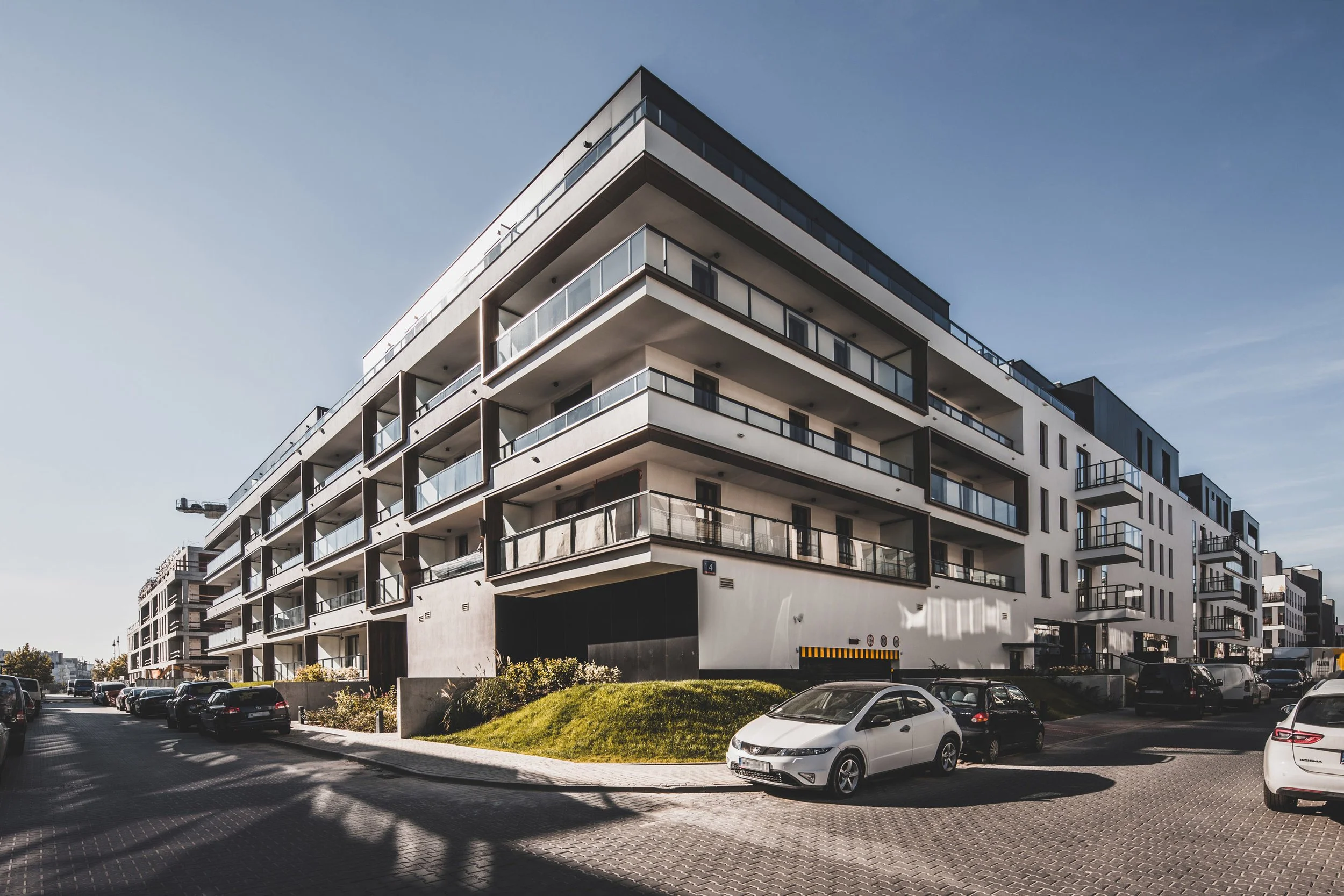 Modern multi-story residential building with balconies, parked cars, and landscaped area in front.