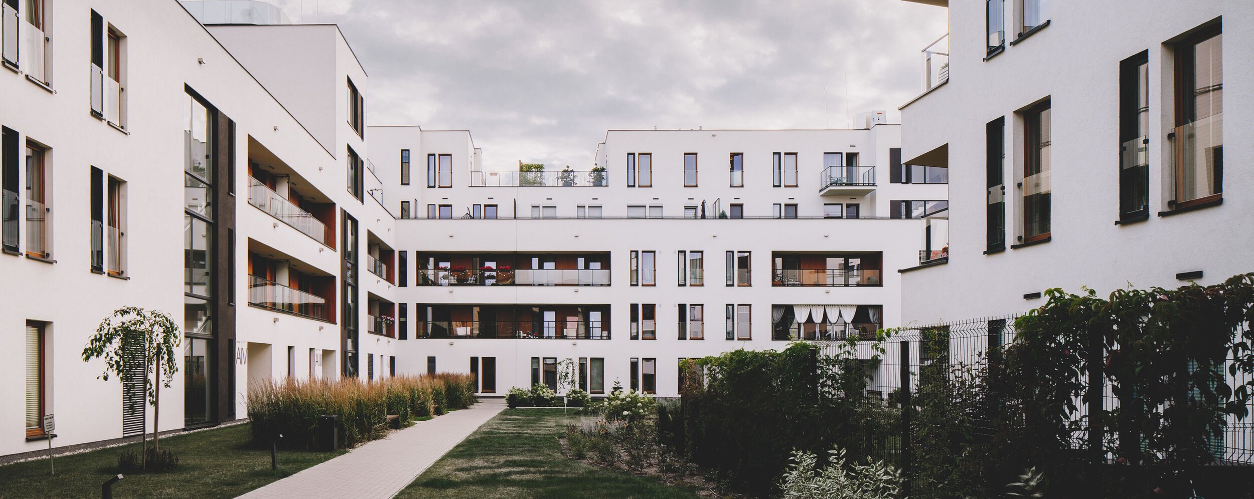 View of modern white apartment buildings with black window frames and balconies, small courtyard with grass and plants, walking path, overcast sky.