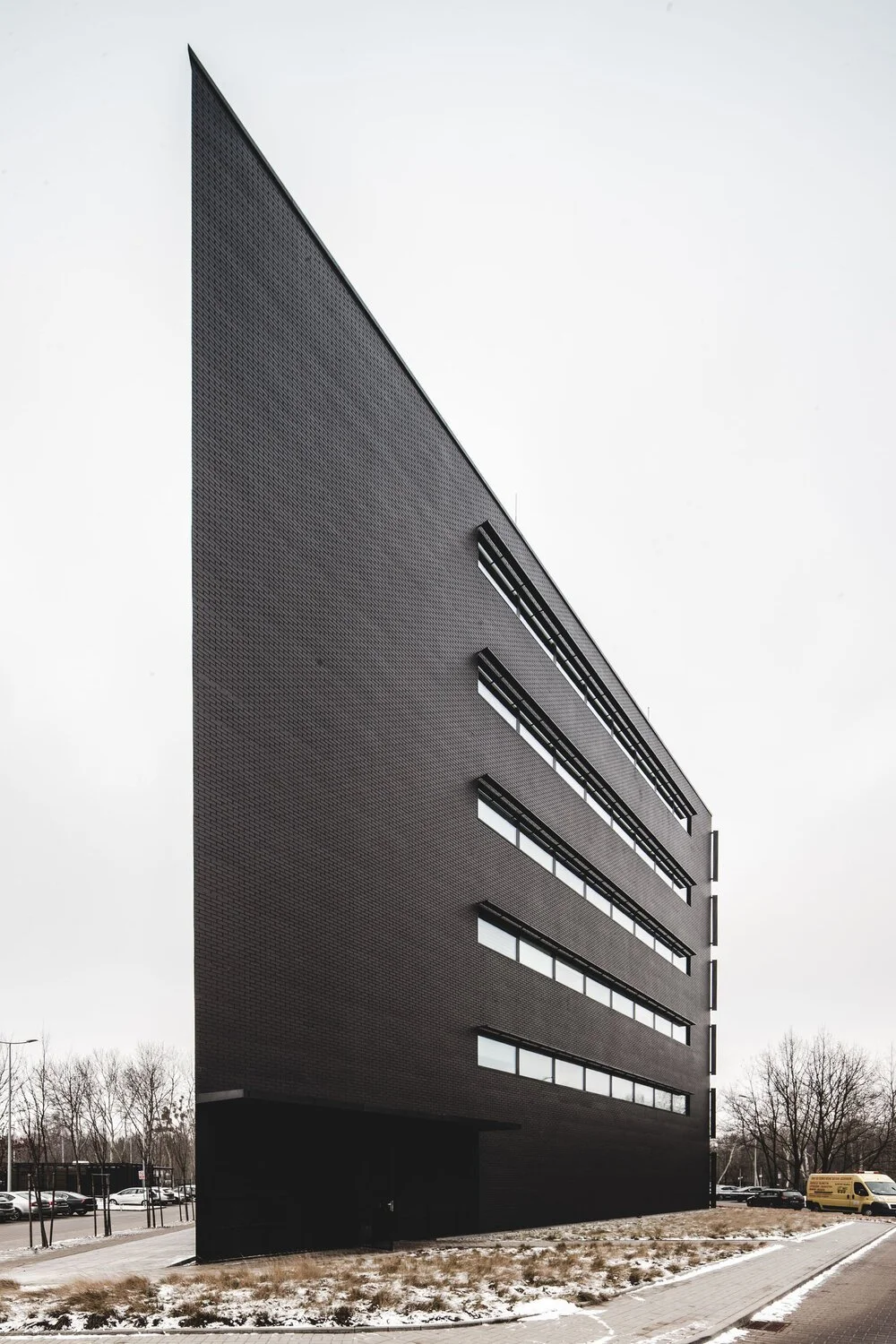 Modern black multi-story building with horizontal windows, surrounded by parking lot and barren trees on overcast day.