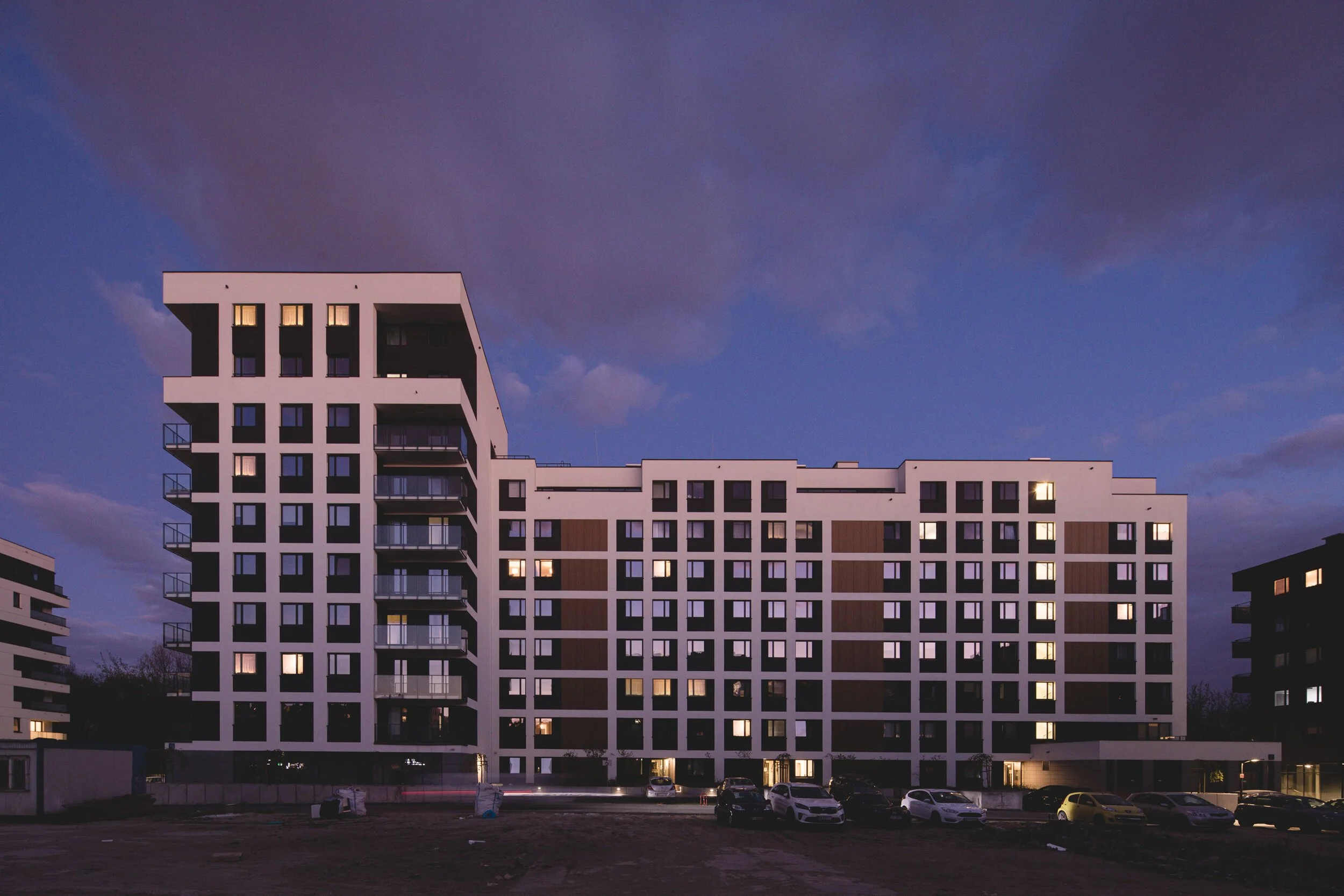 Modern multi-story apartment building at dusk with illuminated windows and a parking lot in the foreground.