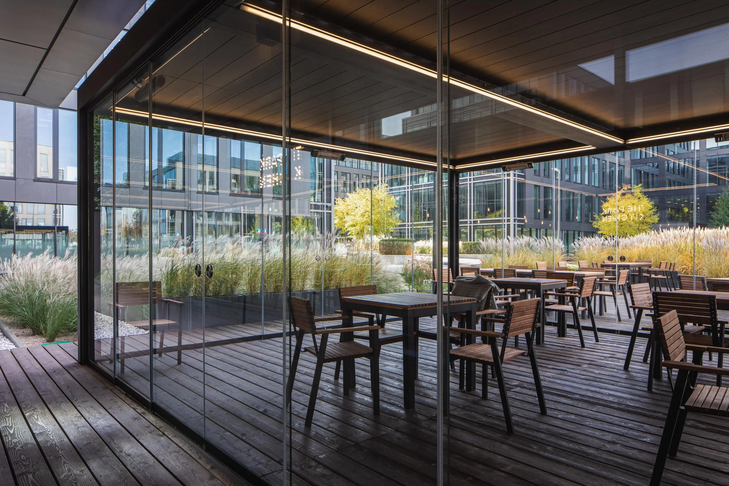 Empty outdoor patio with wooden flooring and tables with chairs, enclosed by glass walls, with modern office buildings and landscaped greenery outside.