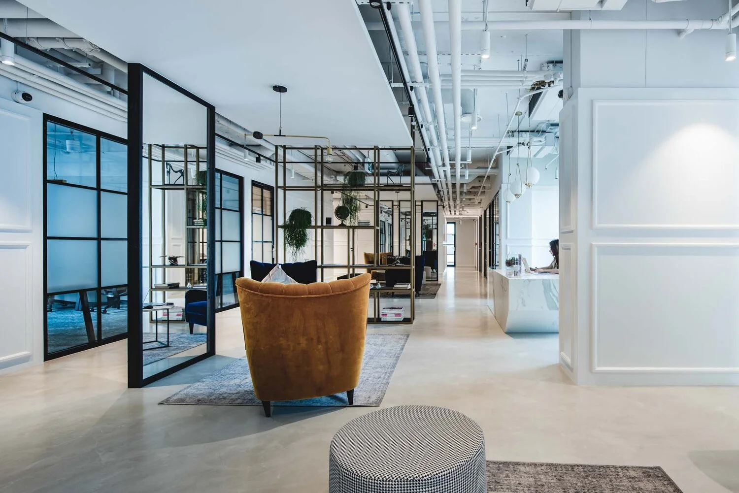 Modern office space with lounge chairs, glass partitions, potted plants, and a reception area with a person working at a desk.