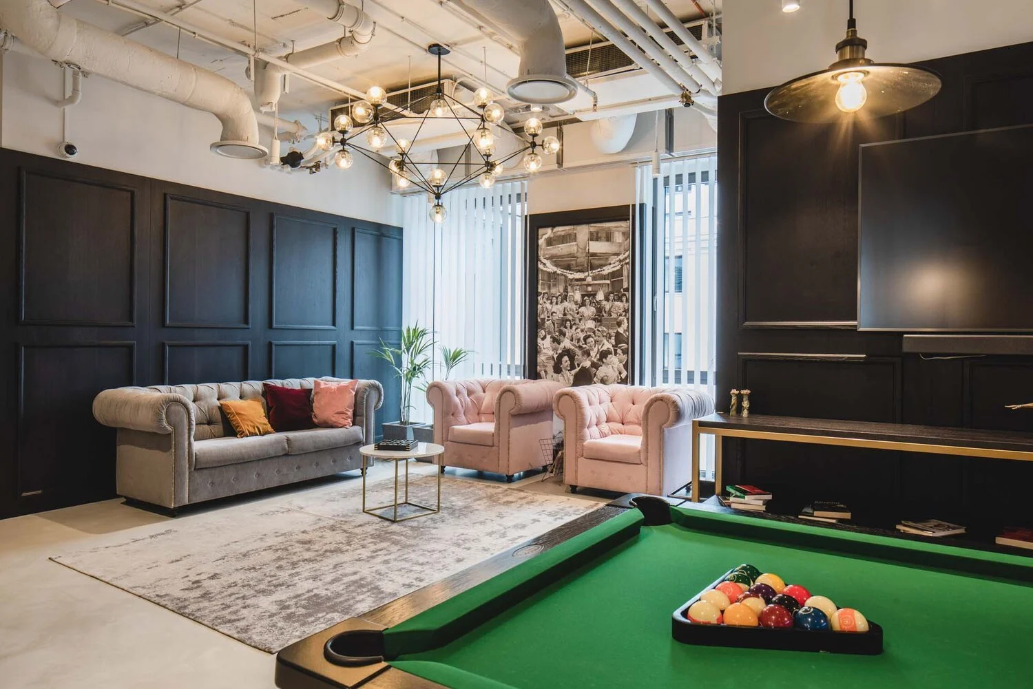 A stylish lounge room with black paneled walls, a gray sofa with colorful cushions, pink armchairs, a pool table with balls, a chandelier, and large windows with vertical blinds.
