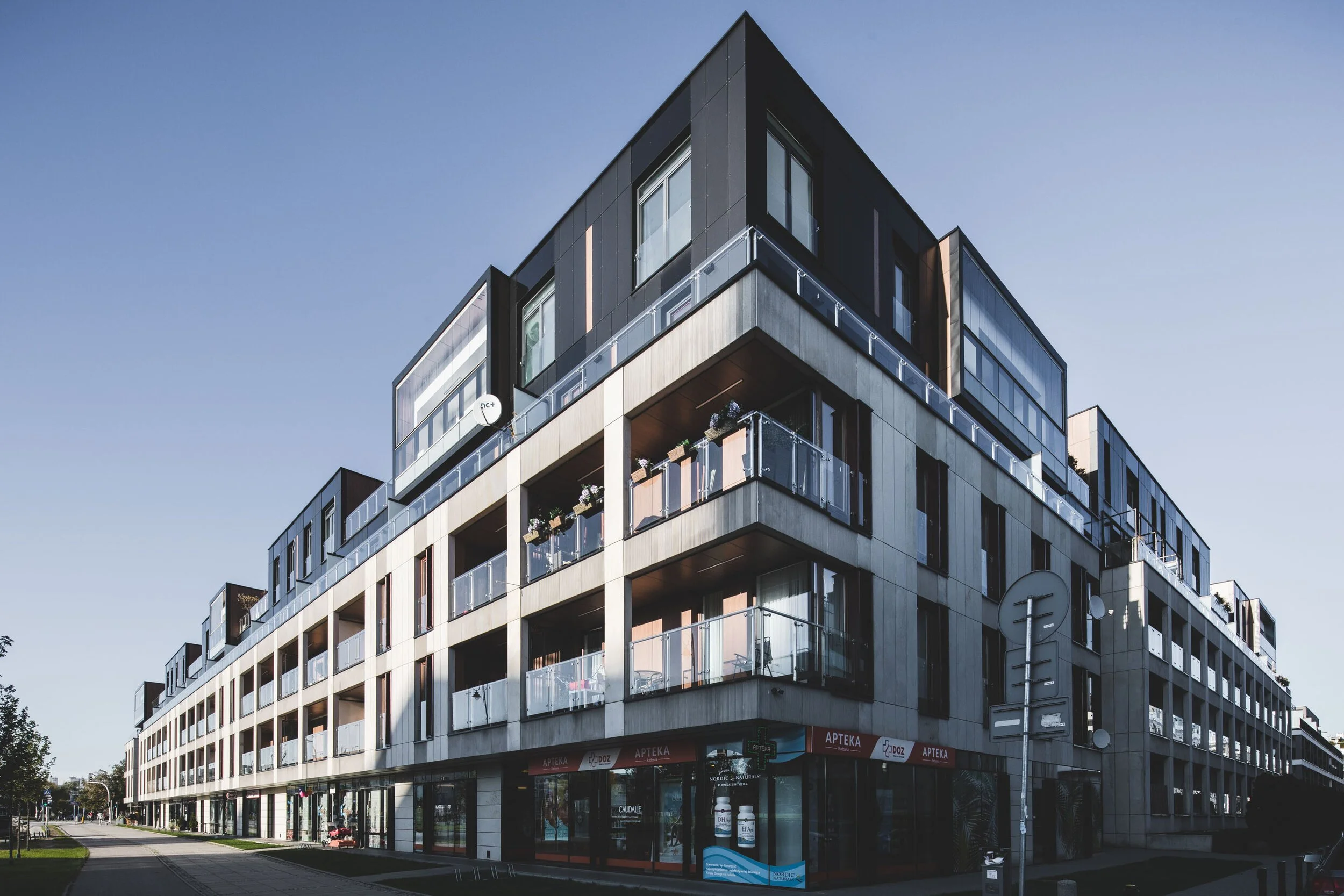 Modern multi-story apartment building with balconies and storefronts at street level, under a blue sky.