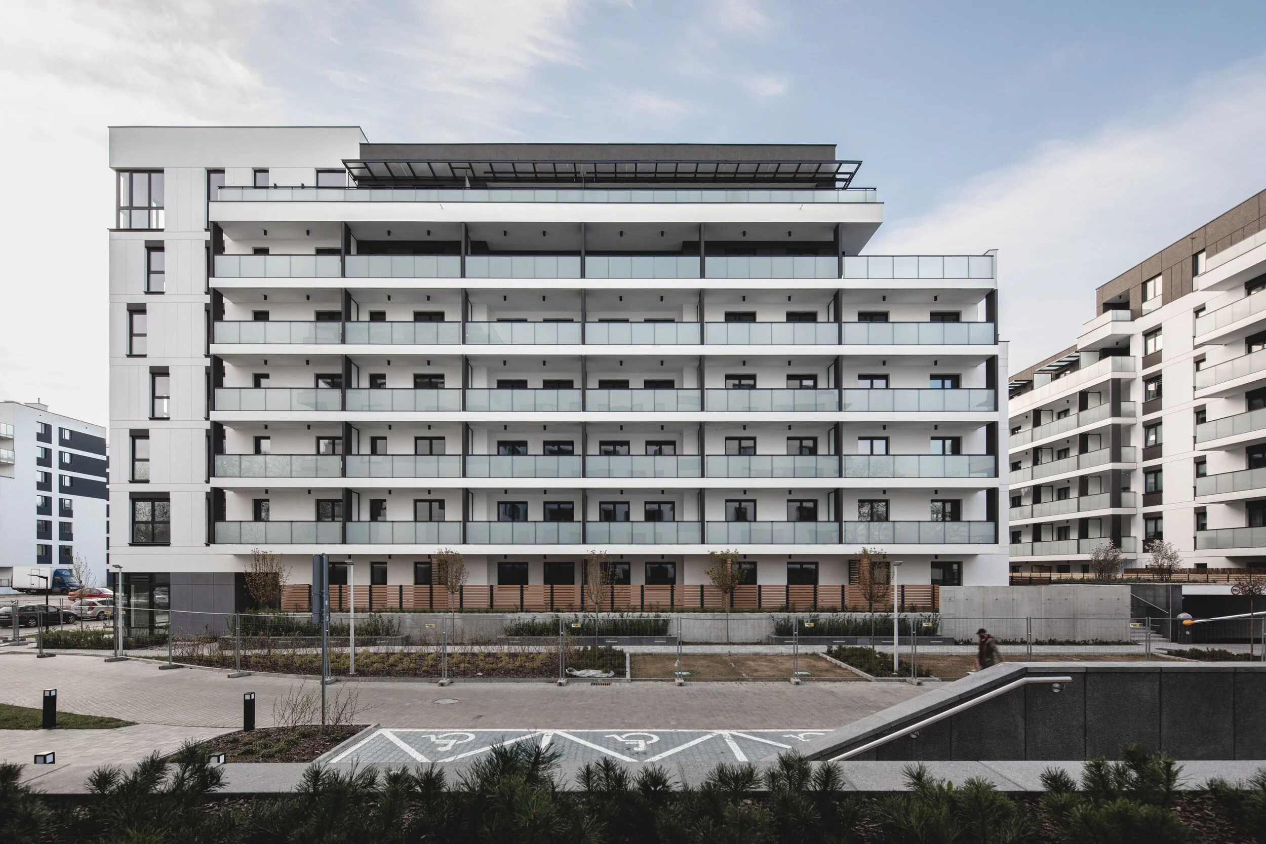 Modern multi-story apartment building with glass balconies, set in an urban environment with landscaped areas and parking spaces in the foreground.