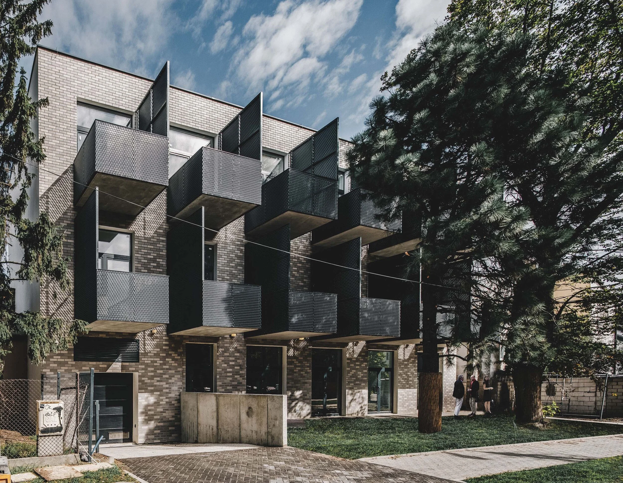 Modern apartment building with black balconies and brick facade, surrounded by trees and a small group of people walking on the sidewalk.
