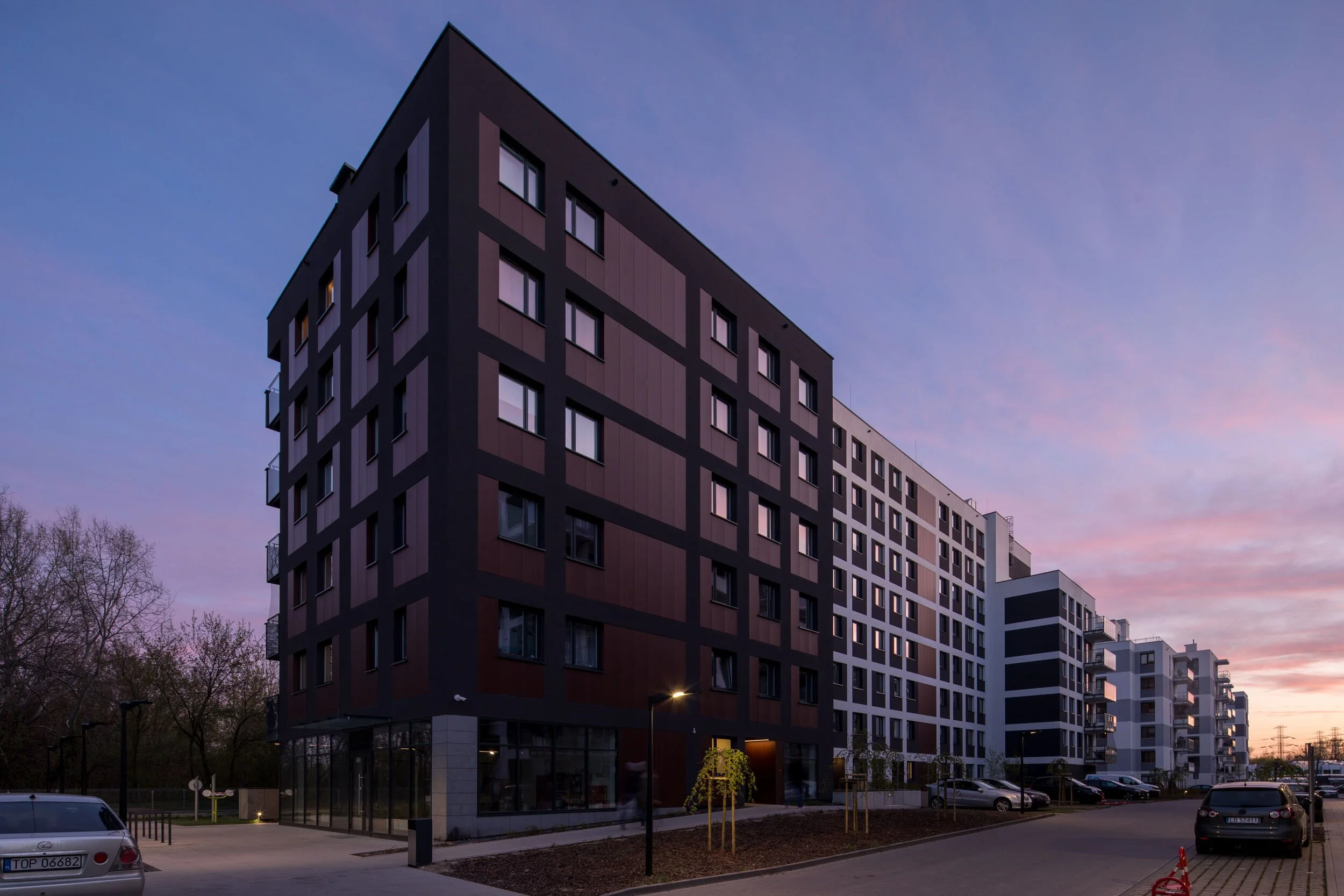 Modern residential apartment building with multiple floors, large windows, and a dark facade, set against a dusk sky with pastel colors. Parking lot and trees in the foreground.