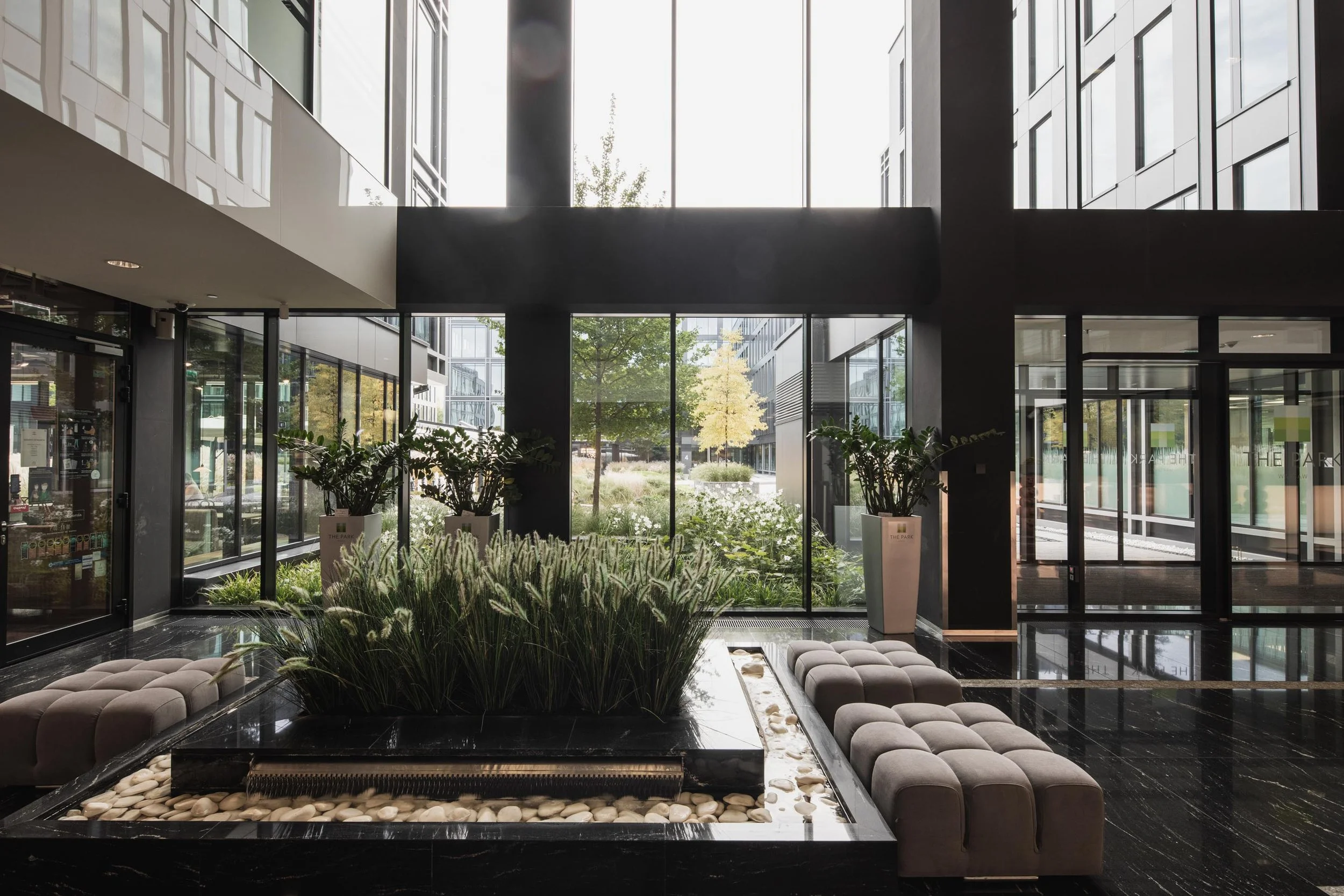 Modern lobby with large glass windows, indoor garden, plants, beige seating, and dark flooring.