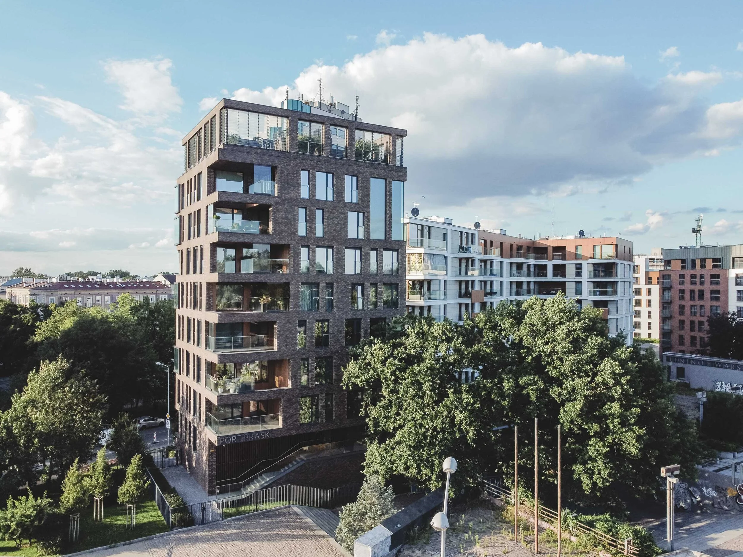 A modern multi-story apartment building with large glass windows and balconies, surrounded by greenery and other residential buildings under a partly cloudy sky.