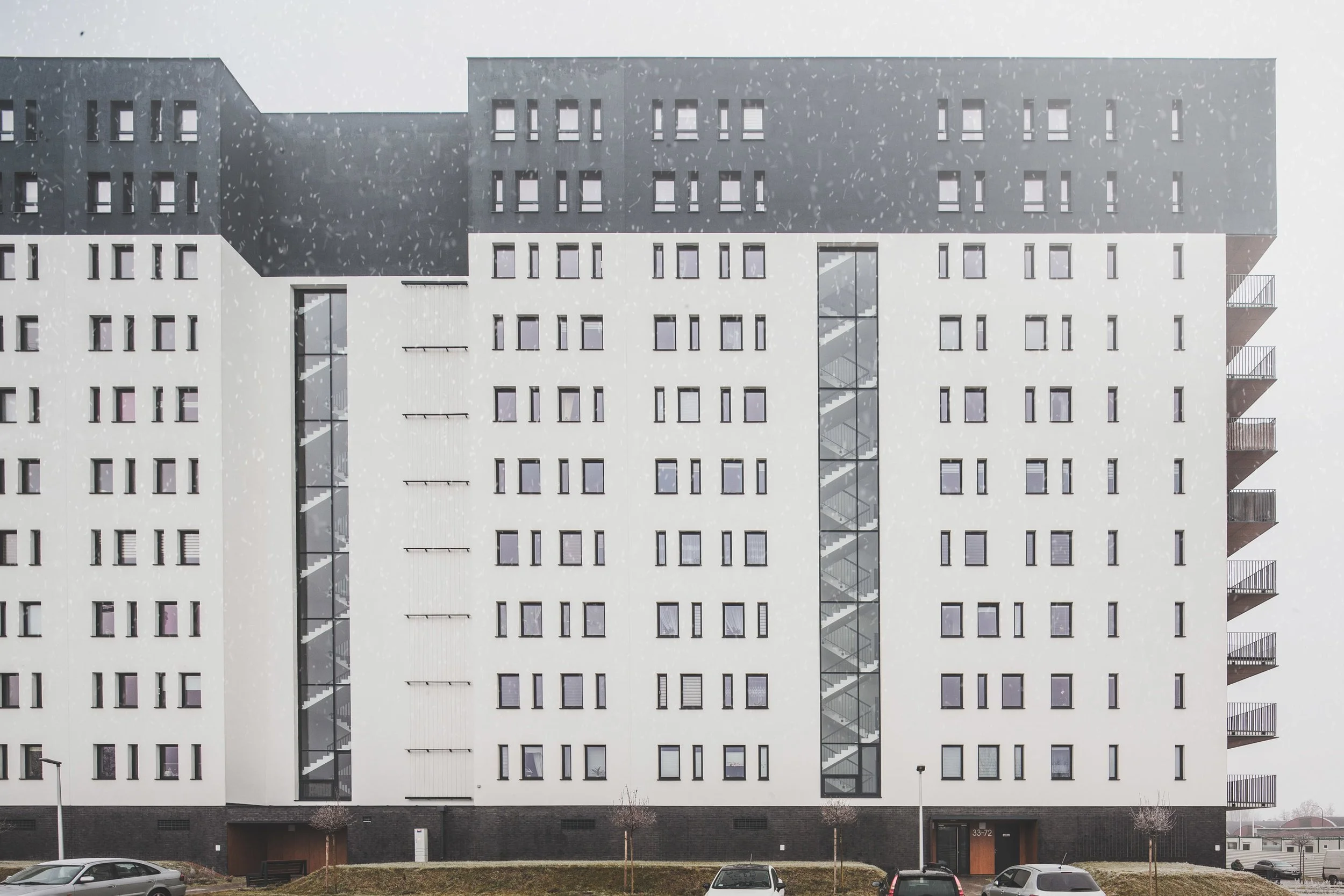 Modern multi-story apartment building with black and white exterior, tall narrow glass stairwell, and balconies on the right side, with cars parked in front and overcast sky in the background.